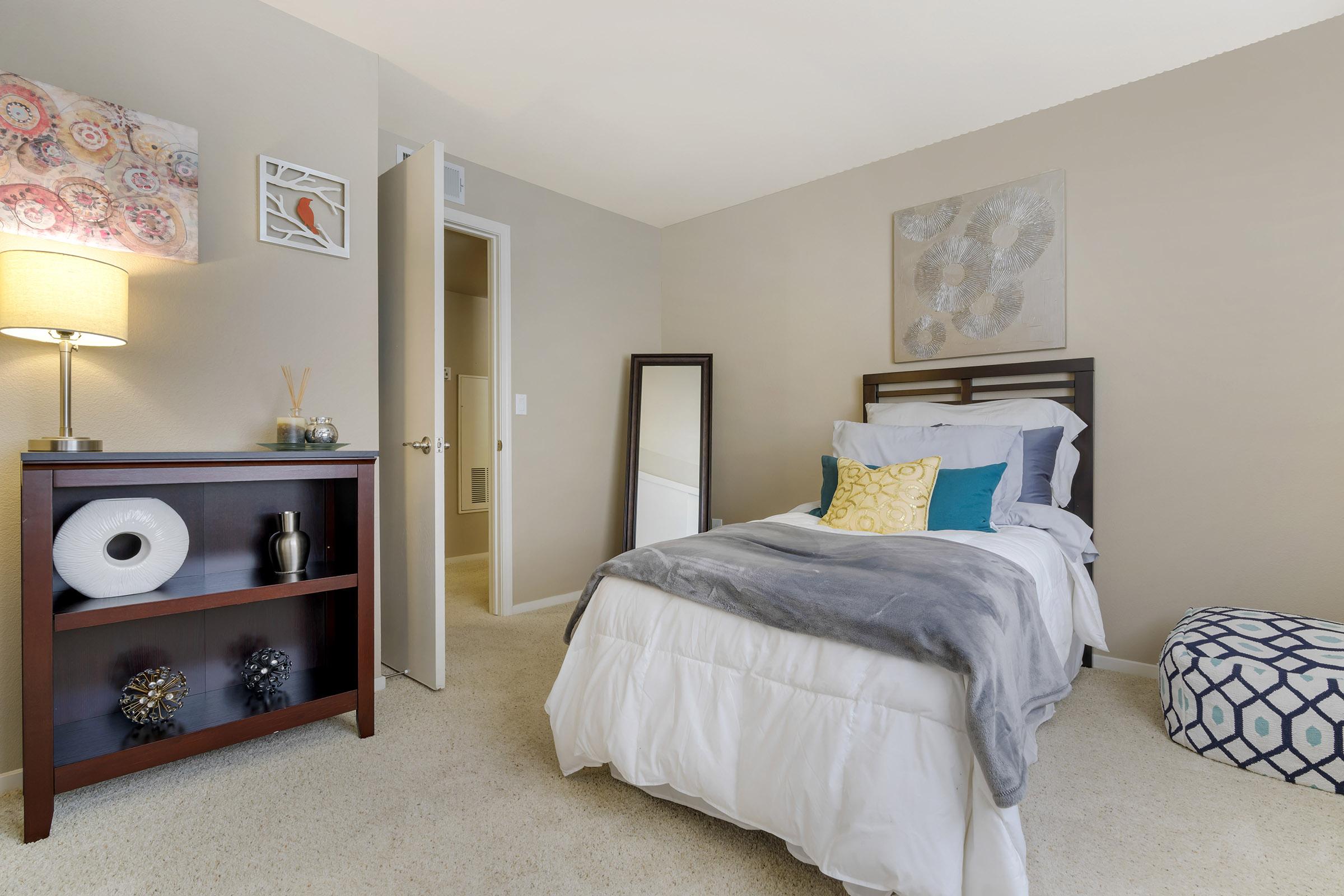 A cozy bedroom featuring a single bed with a grey throw, decorative pillows, and a colorful wall art. There is a bedside lamp, a mirror, a bookshelf with decorative items, and a patterned ottoman. The room has light-colored walls and carpet, creating a warm and inviting atmosphere.