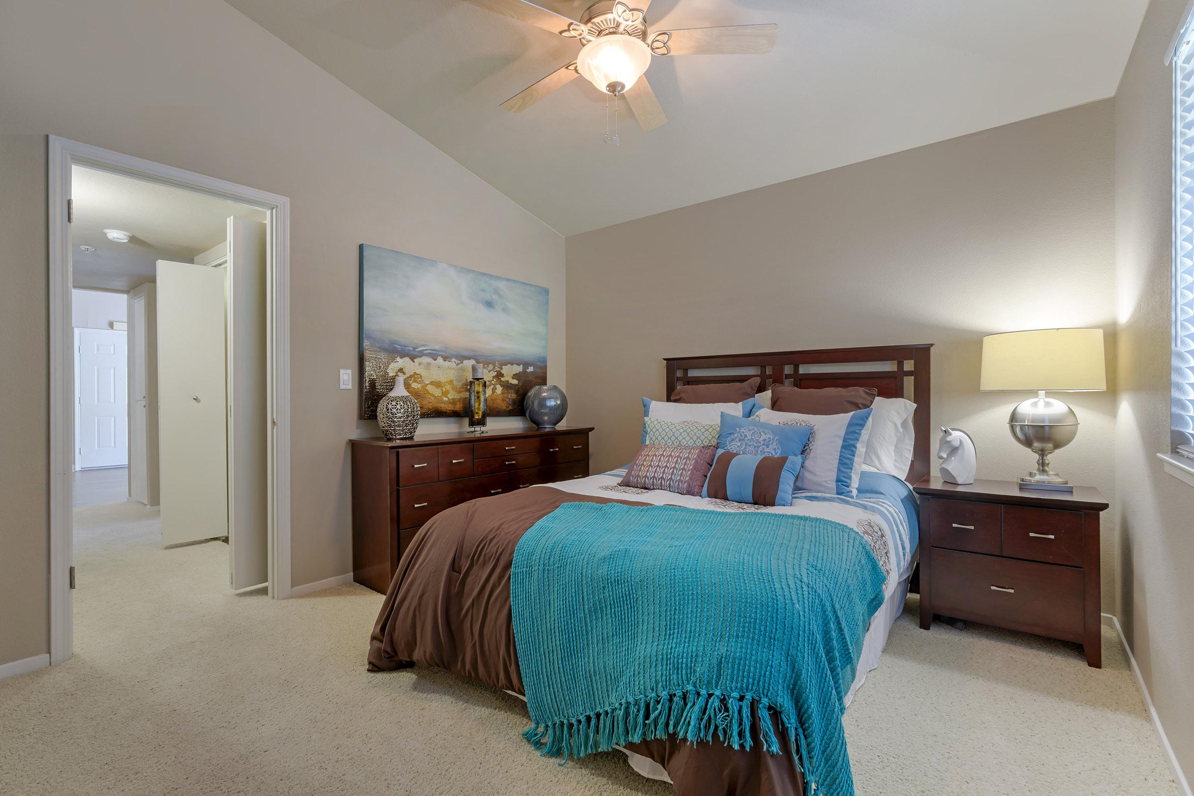 A cozy bedroom featuring a neatly made bed with a turquoise throw blanket and decorative pillows. There are two nightstands with lamps on either side, a dresser adorned with decorative vases, and a large piece of artwork above the bed. Soft light enters through a window, enhancing the warm atmosphere.