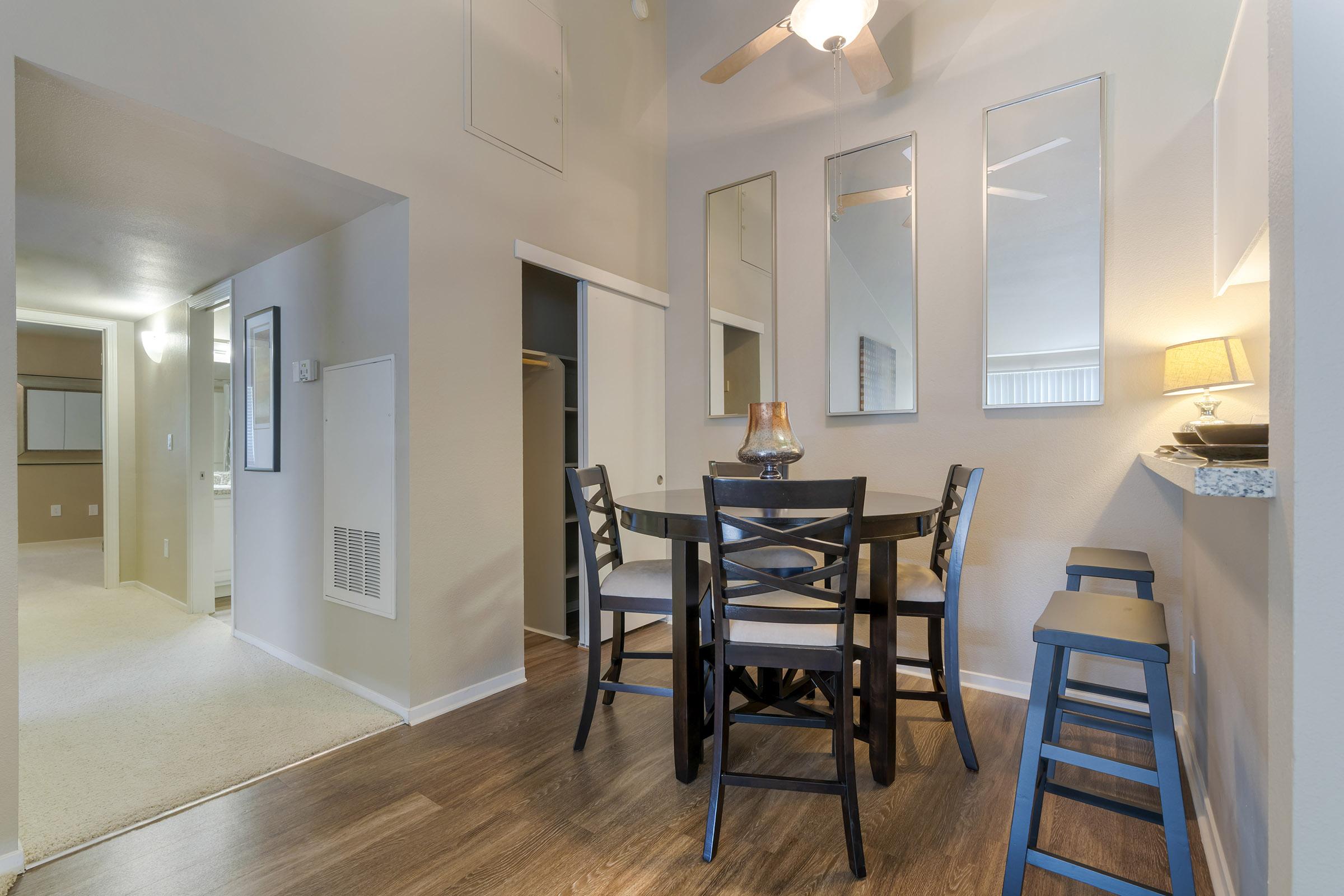 A modern dining area featuring a round black table with four chairs, surrounded by mirrors on the wall. A lamp sits on the table, and there's a bar area with two blue stools. The space has warm wood flooring and soft lighting, creating an inviting atmosphere. Doorways lead to a hallway and additional rooms.