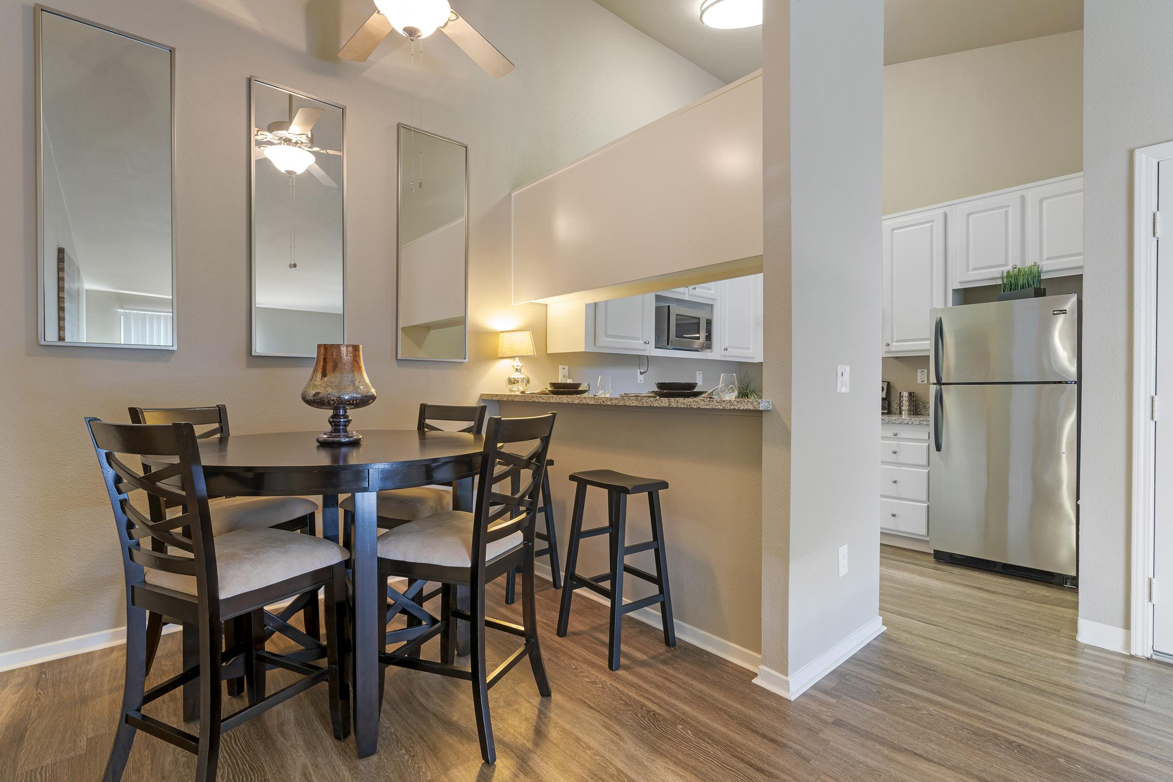 A modern kitchen and dining area featuring a round black table with four chairs, a small lamp on the table, and a countertop with bar stools. The kitchen has white cabinets, a stainless steel refrigerator, and mirrors on the wall, contributing to a bright and inviting atmosphere.