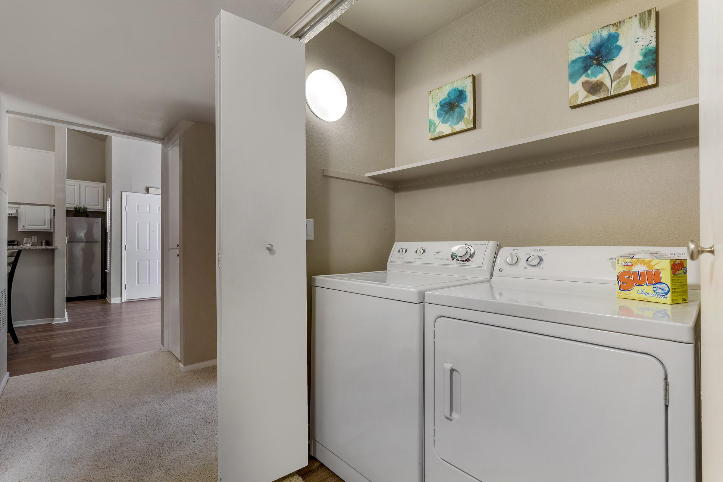 A laundry room featuring a white washer and dryer, with a shelf above and two decorative floral prints on the wall. A round window lets in natural light. The room has a beige wall color and a doorway leading to another area of the home that is partially visible.