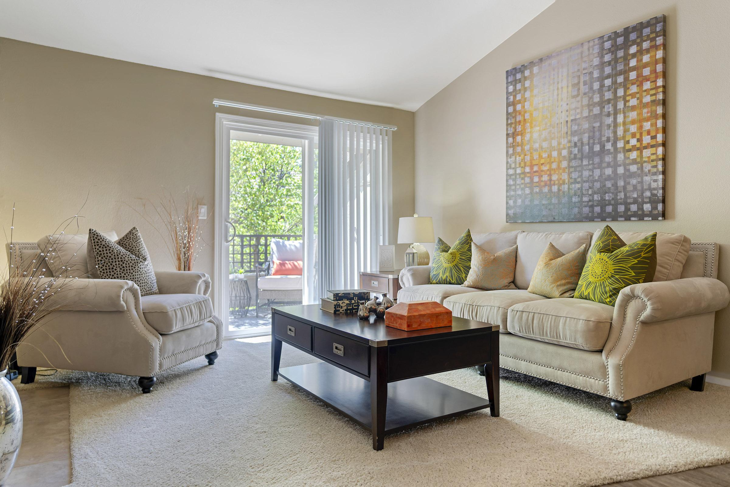 A cozy living room featuring a light-colored sofa with patterned cushions, a beige armchair, and a dark wooden coffee table with decorative items. Natural light fills the space, highlighting the textured wall art and a view of greenery through sliding glass doors. A soft area rug adds warmth to the scene.