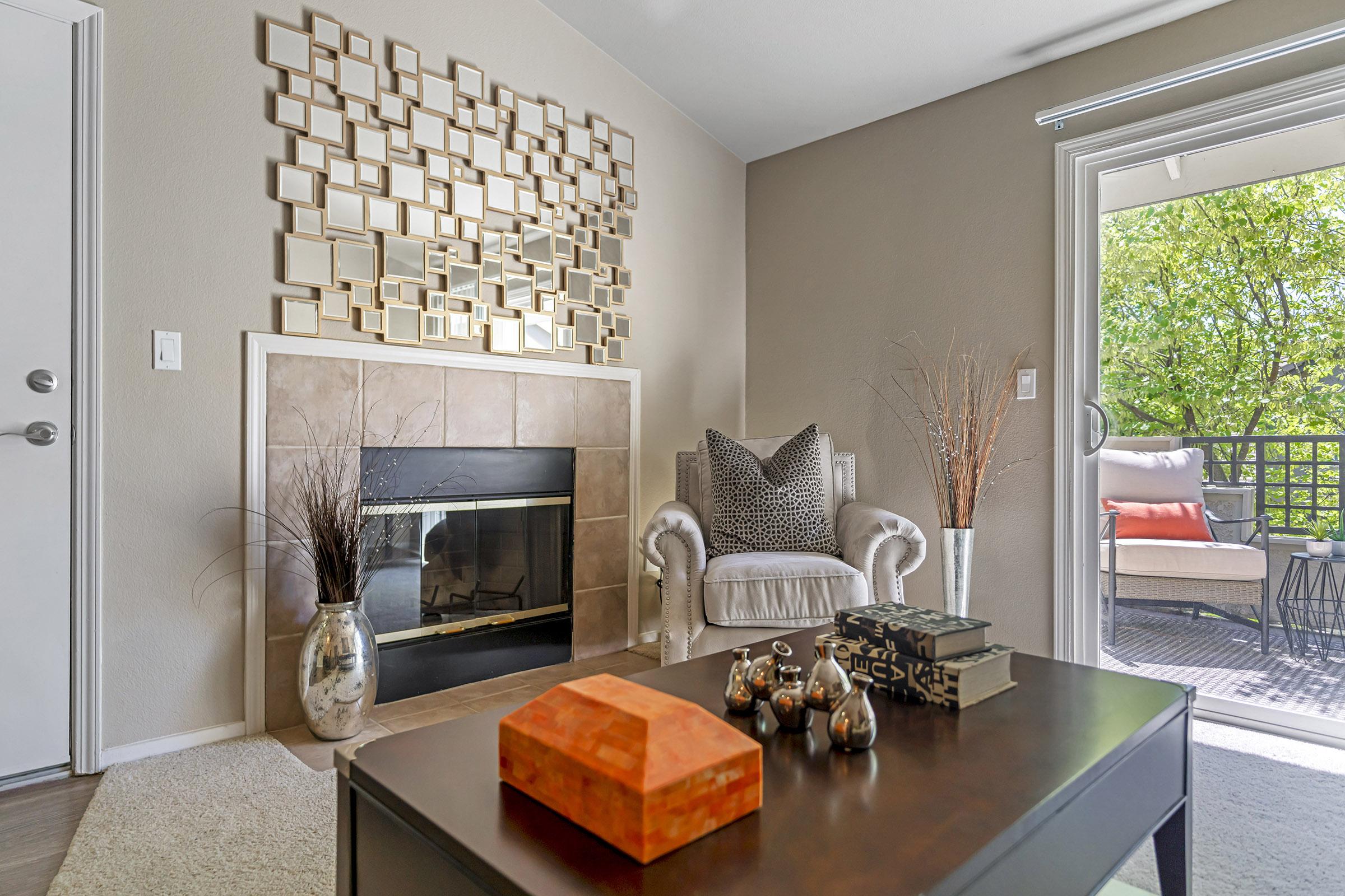 A cozy living room featuring a light-colored armchair with patterned cushions, a fireplace with a decorative mirror above it, and a modern coffee table. The decor includes vases and a wooden box, with a sliding glass door leading to a balcony, showcasing greenery outside. Soft lighting enhances the inviting atmosphere.