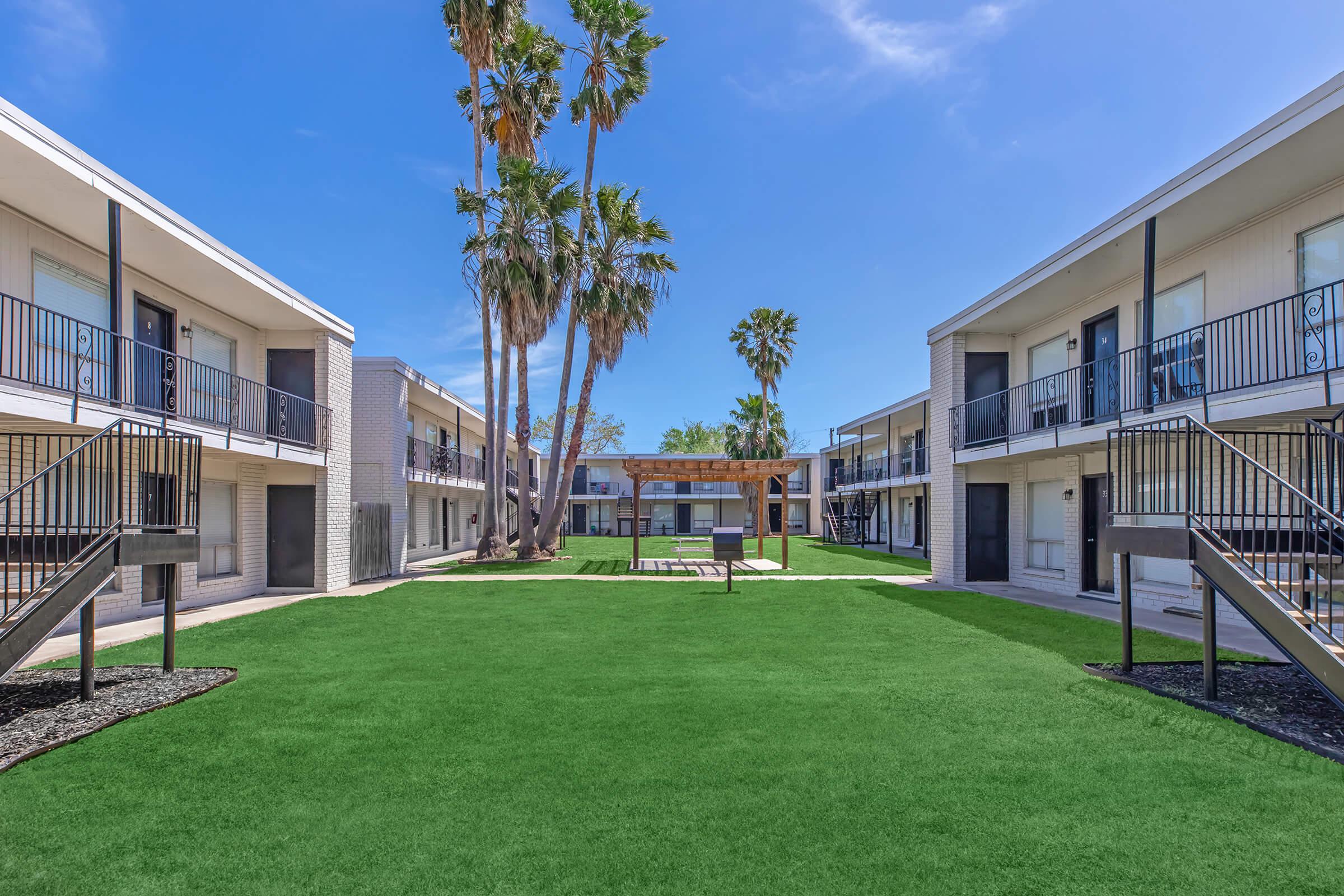 View of an apartment complex courtyard featuring two two-story buildings with balconies, surrounded by palm trees. A grassy lawn sits in the center, complemented by a wooden pergola and lounge chairs, creating a relaxing outdoor space under a clear blue sky.