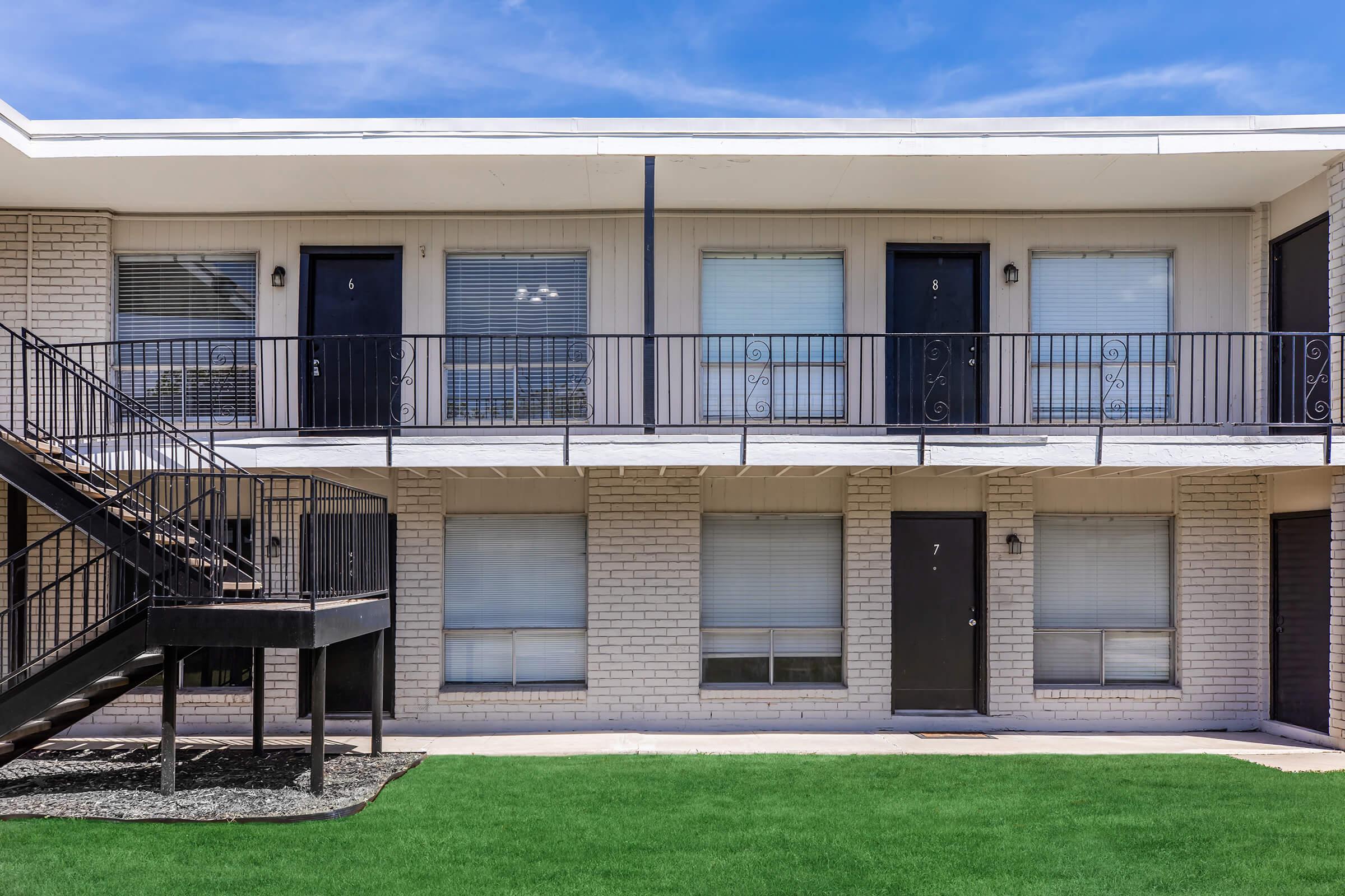 A two-story apartment complex featuring several units with black doors and white brick exterior. The ground level has a stairway on the left, leading to the upper floor. The grassy area in front is neatly maintained, and the sky above is clear with blue tones.