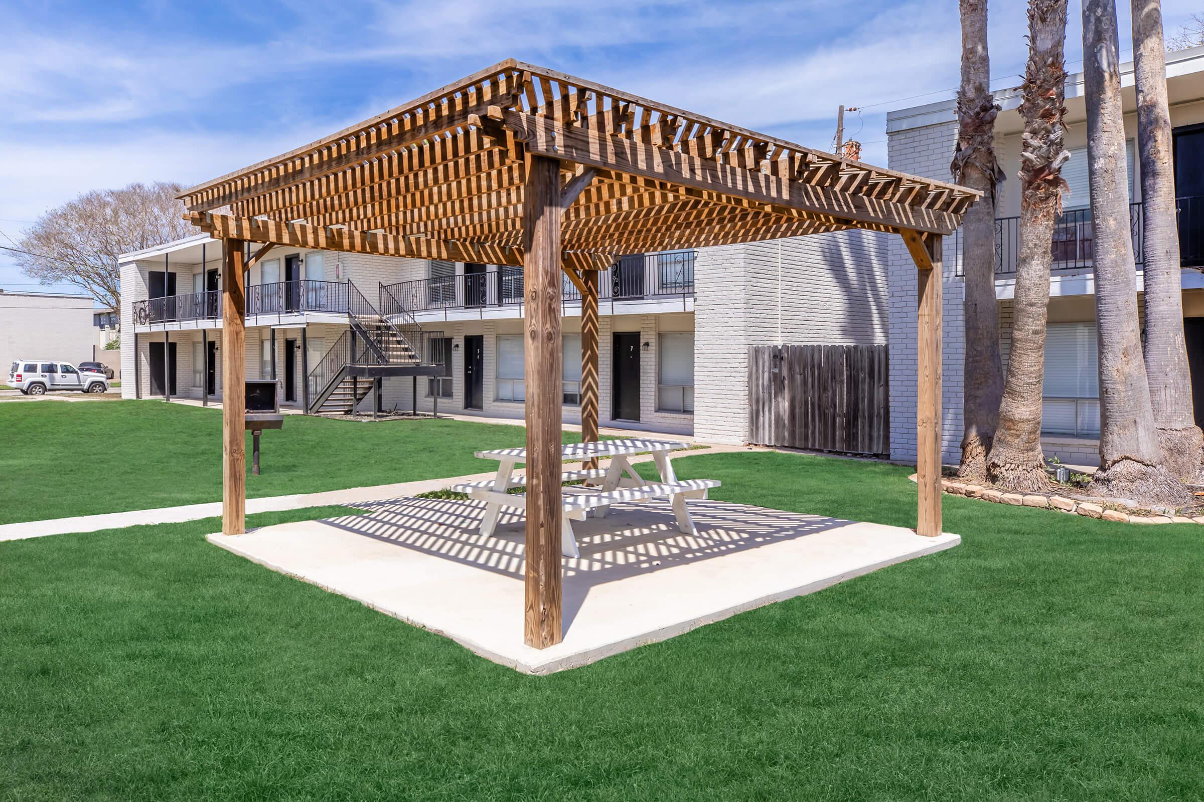 A wooden pergola over a picnic table on a shaded patio, surrounded by green grass. In the background, there are multi-story apartment buildings with balconies and staircases. Bright blue sky with clouds overhead.