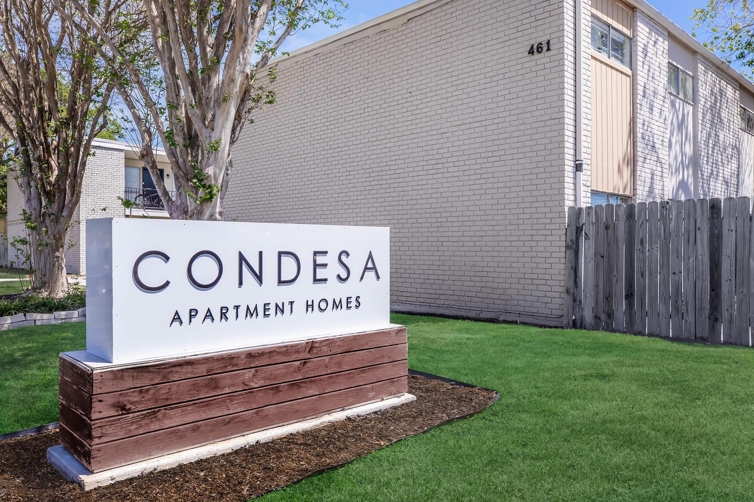 Sign for Condesa Apartment Homes located at 461, featuring a modern design with a white background and bold lettering. The sign is situated in a well-maintained green area with trees and a wooden fence in the background.