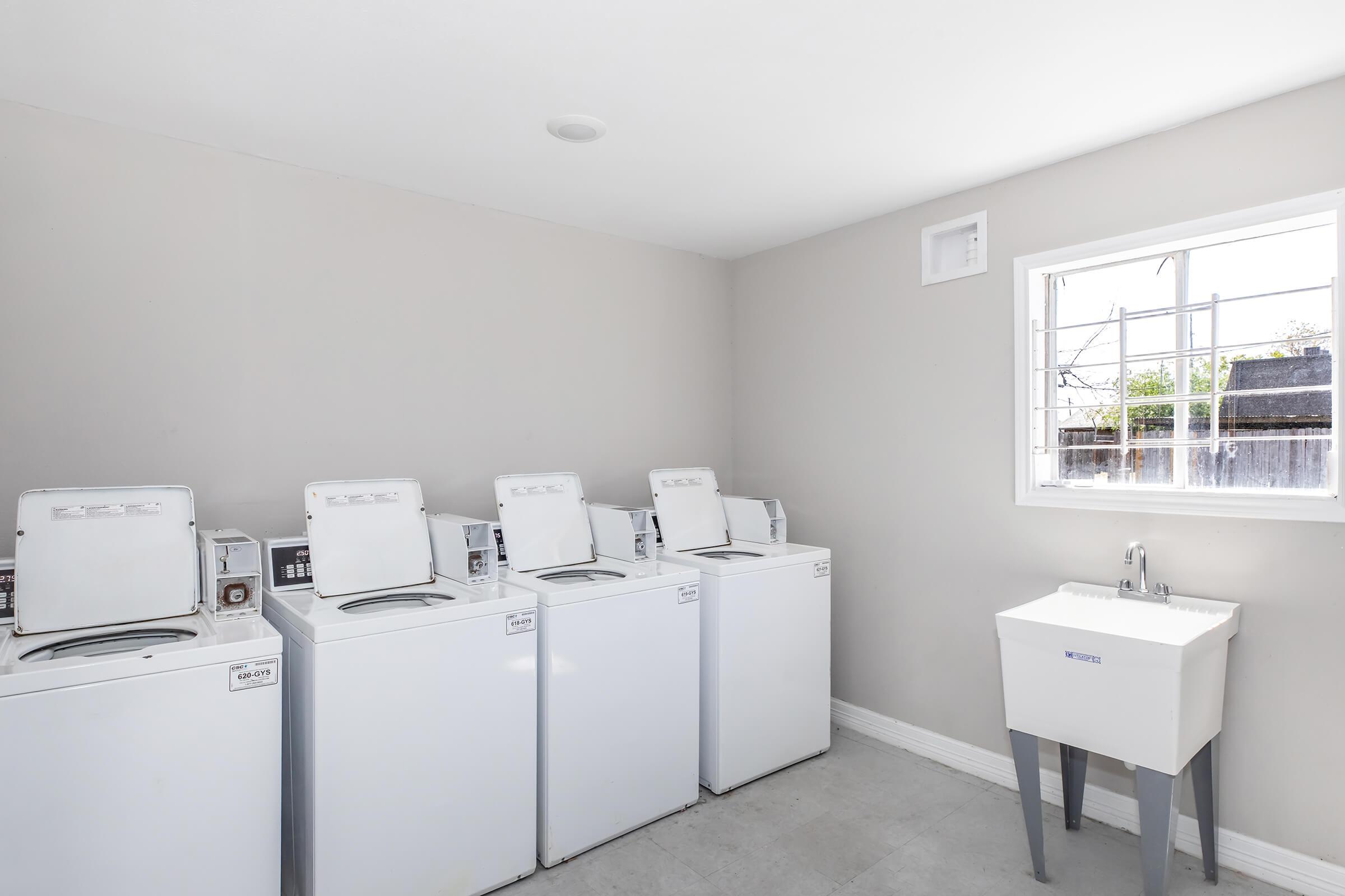 A clean and organized laundry room featuring four white washing machines aligned against a light grey wall. A small sink stands to the right, and a window above provides natural light, illuminating the space. The floor is light-colored, enhancing the room's bright and airy feel.