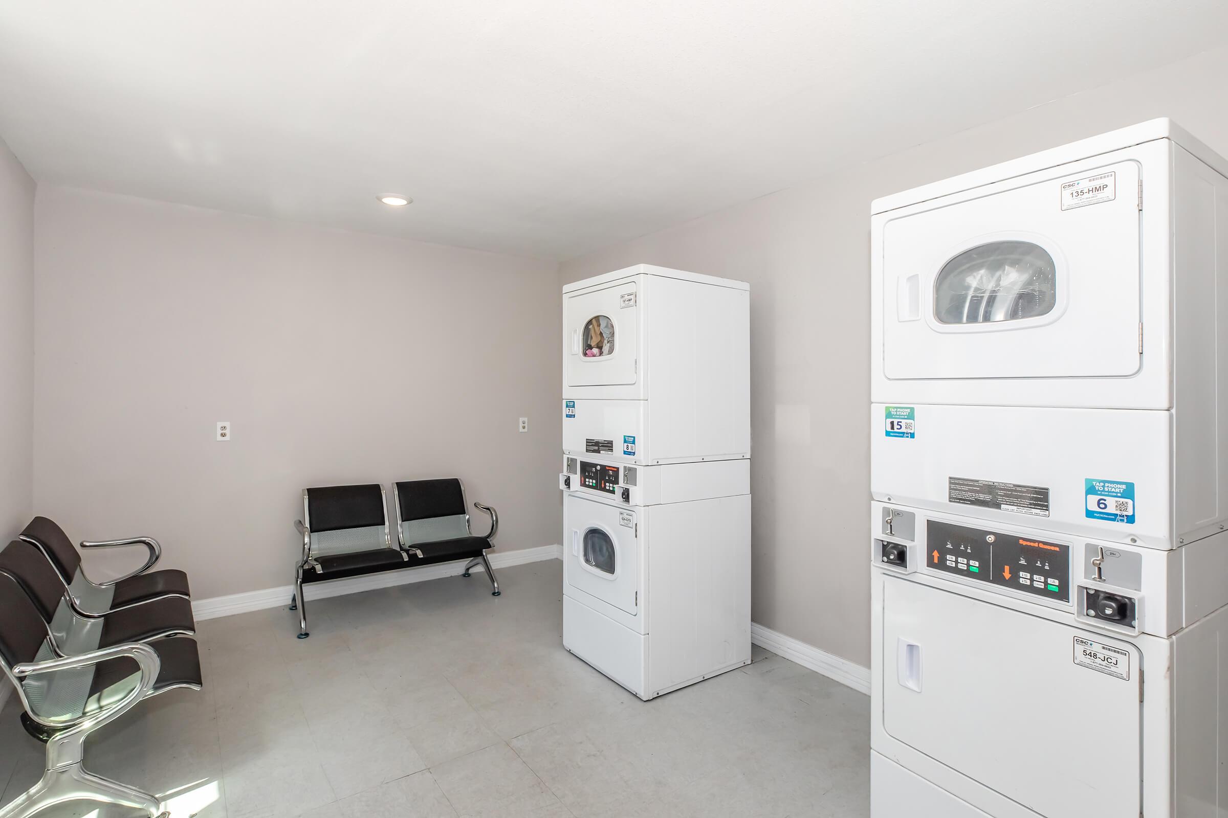 A clean, well-lit laundry room featuring two stacked washer-dryer units and two modern benches for seating. The walls are painted a neutral color, and the flooring is light-colored tile. The space appears tidy and organized, suitable for residents to do laundry comfortably.