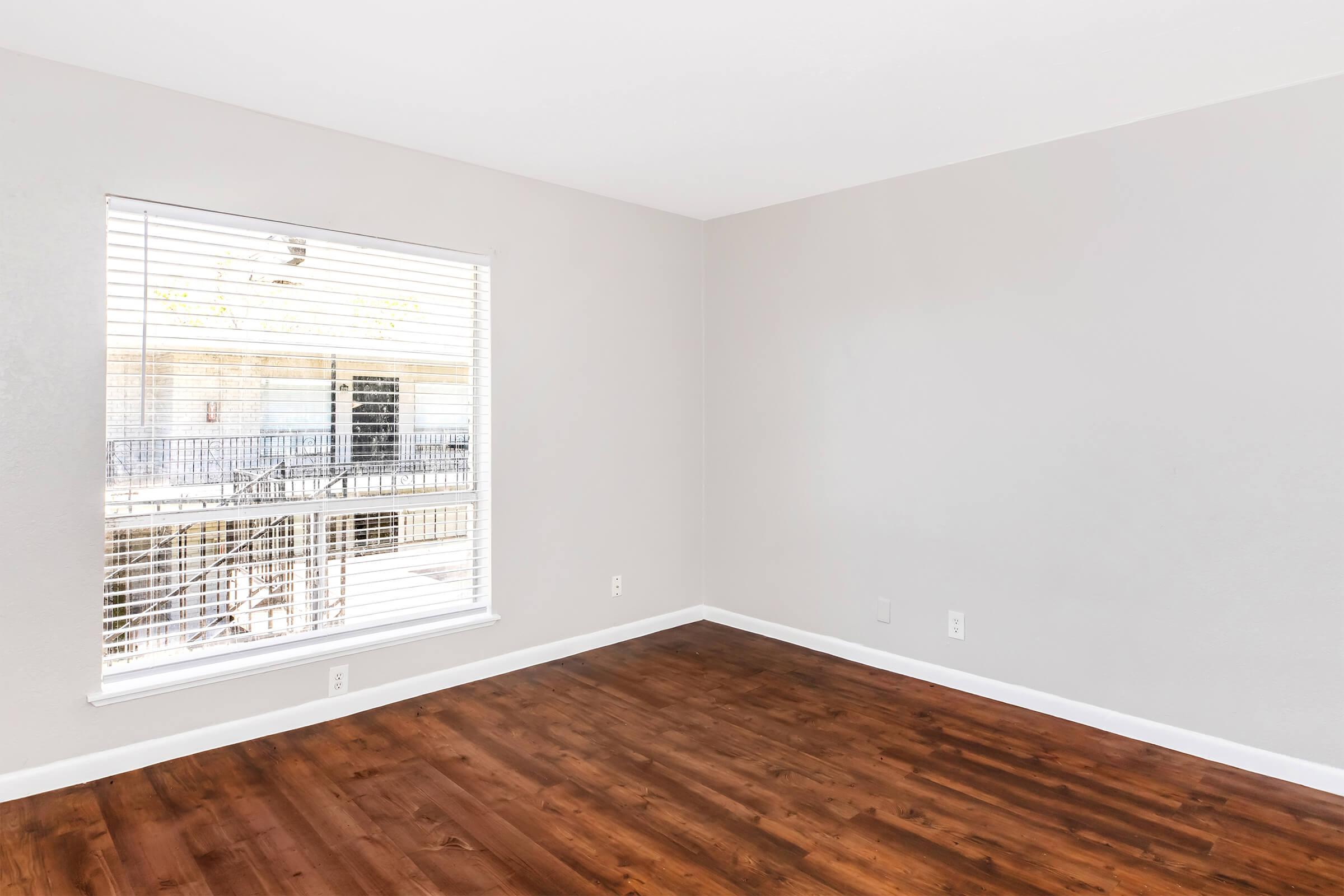 Interior view of a vacant room featuring light gray walls, a large window with white blinds, and a wooden floor. The window displays an outdoor balcony area. The room has no furniture, providing a spacious and minimalistic atmosphere.