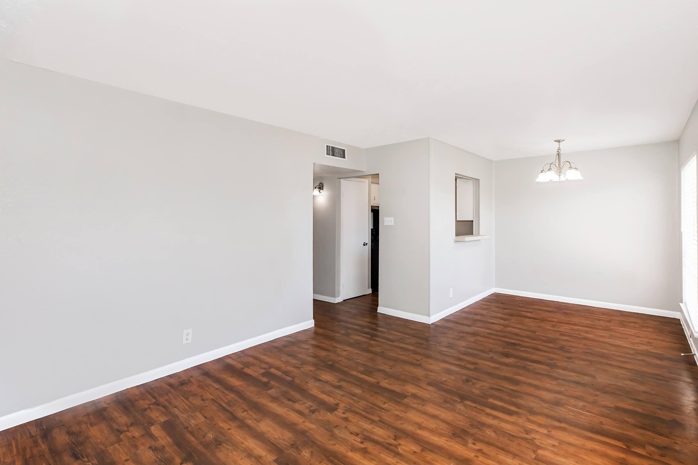 A spacious interior room featuring light gray walls, hardwood-style flooring, and a bright atmosphere. There's a doorway leading to another room on the left, and a small dining area with three hanging lights in the corner, providing a functional and inviting living space.