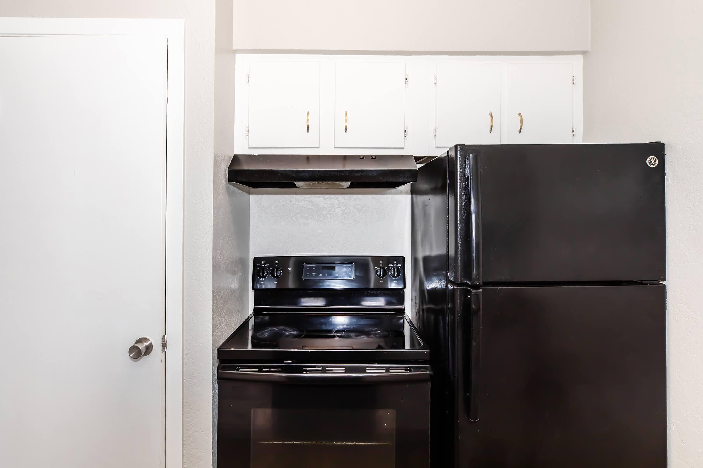 A compact kitchen space featuring a black stove and refrigerator. The stove is situated between the fridge and white cabinetry above. The walls are painted a light color, and a door is visible to the left, suggesting a small, functional area ideal for cooking and storage.