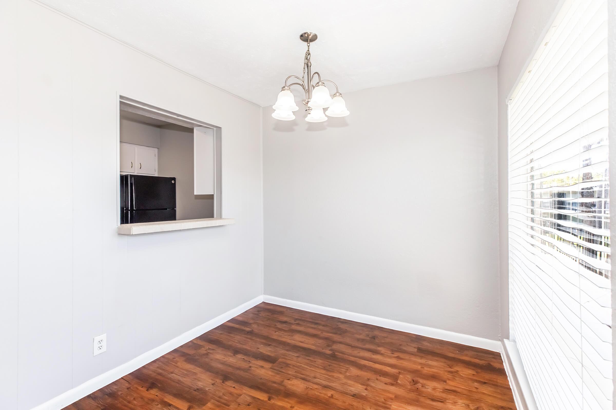 A bright, minimalistic dining area featuring a modern chandelier with four light fixtures. There is an open window with blinds allowing natural light, and a counter opening leading to a kitchen area. The floor is a warm hardwood finish, and the walls are painted in a light gray color.