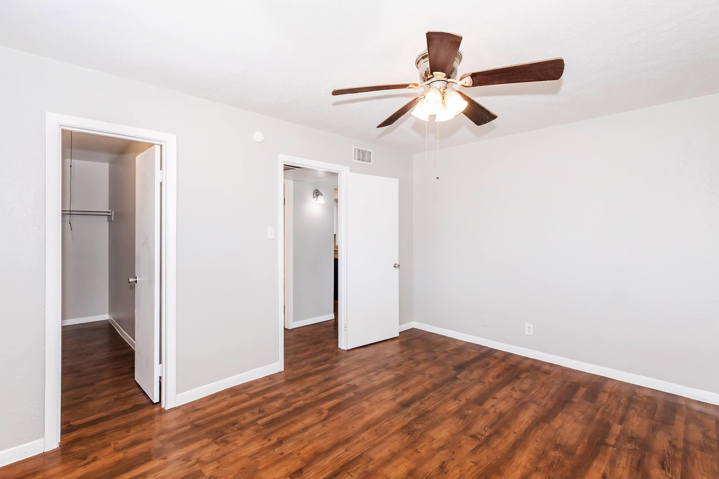 A well-lit, empty room featuring a ceiling fan, hardwood-style flooring, and light gray walls. A closet is visible through an open door on the left, with another door leading to a separate area. The space is uncluttered and ready for furnishing.