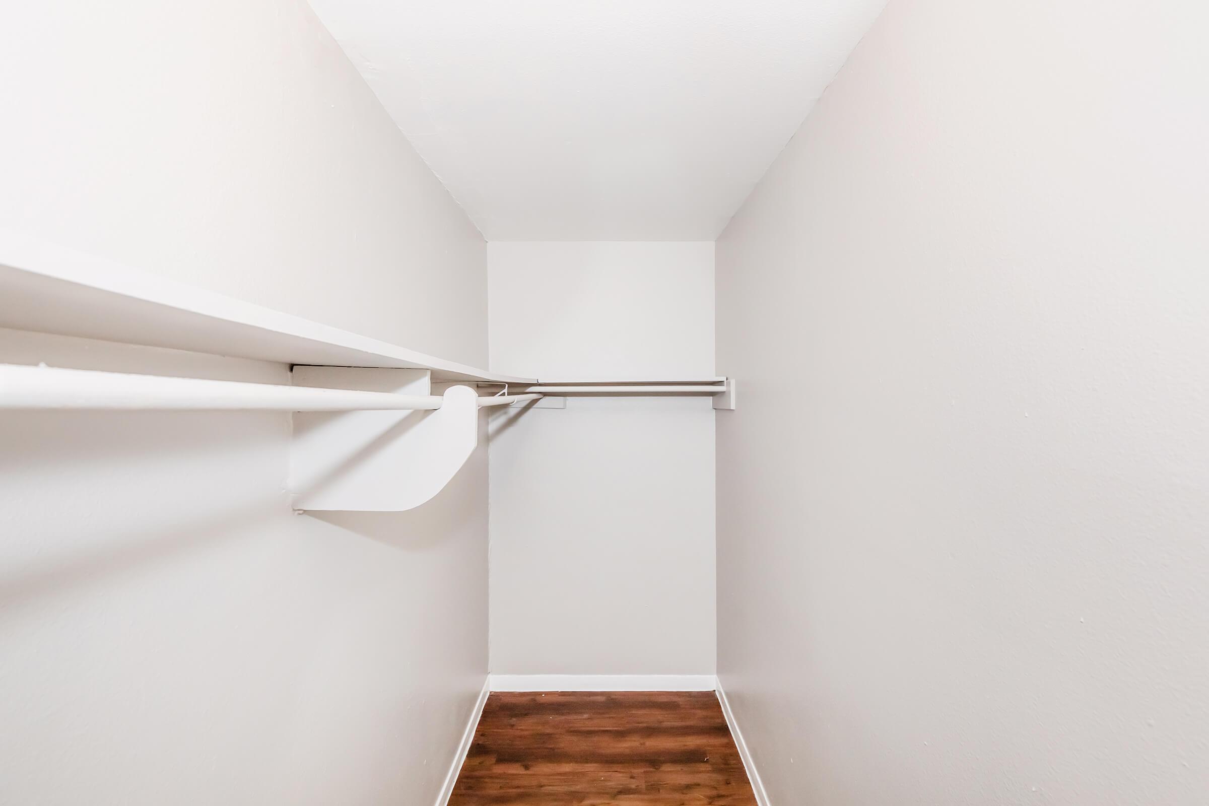 Empty closet with white walls and a wooden floor. Two white hanging rods run parallel along the walls, creating an open and spacious feel. The simplicity of the design enhances its functionality, making it suitable for storage or organization.