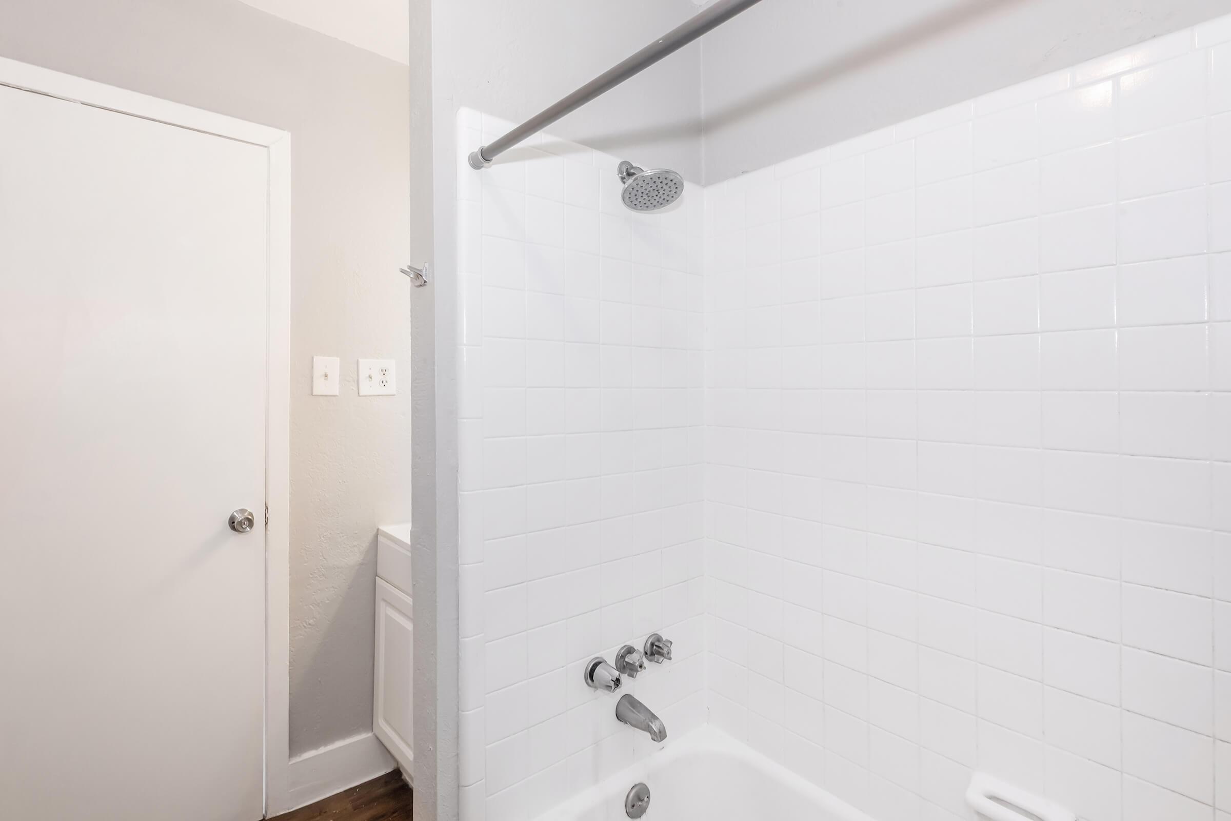 A bright, clean bathroom featuring a white tiled bathtub with a showerhead and chrome fixtures. To the side, there is a closed white door, and light gray walls complete the modern look. The floor has a warm wooden texture, adding a touch of warmth to the space.