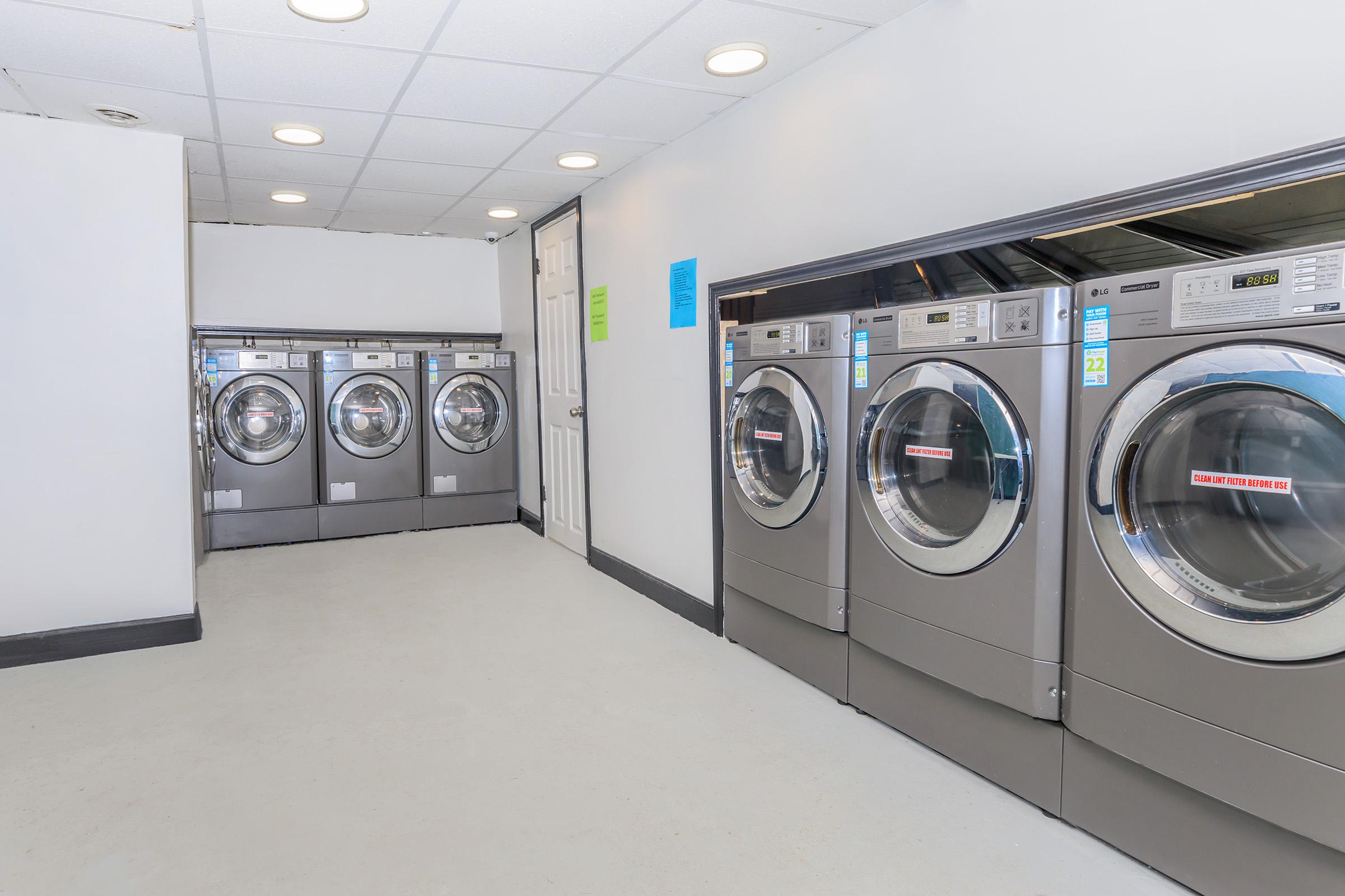 A clean and bright laundry room featuring several commercial washing machines and dryers aligned against the wall. The floor is light-colored, and there are signs on the walls, likely indicating instructions or policies. The space appears well-maintained and organized for users.
