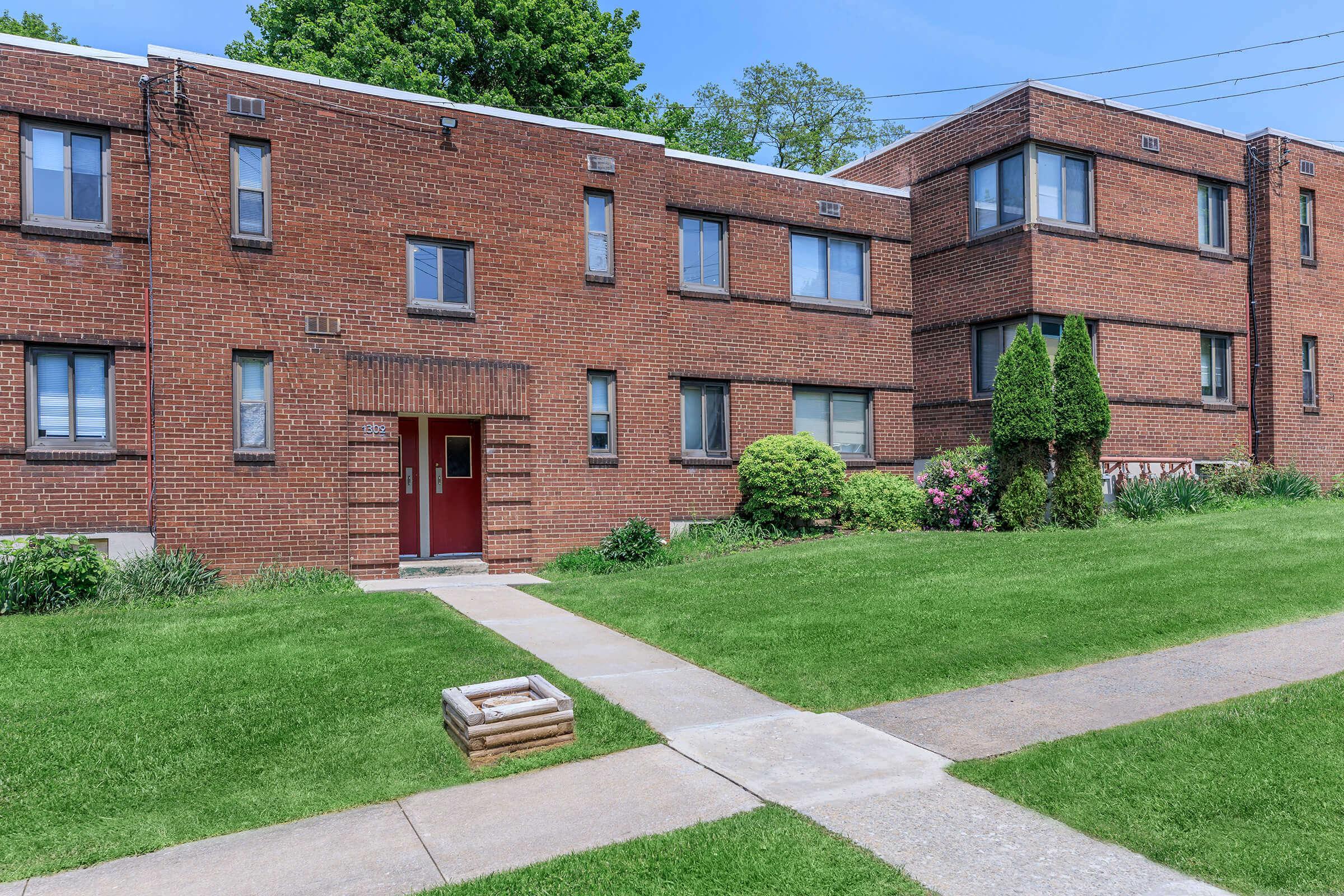 A red brick apartment building featuring multiple windows and a red entrance door, surrounded by a green lawn and landscaped shrubs. A concrete pathway leads to the entrance, and the setting includes a clear blue sky with trees in the background.