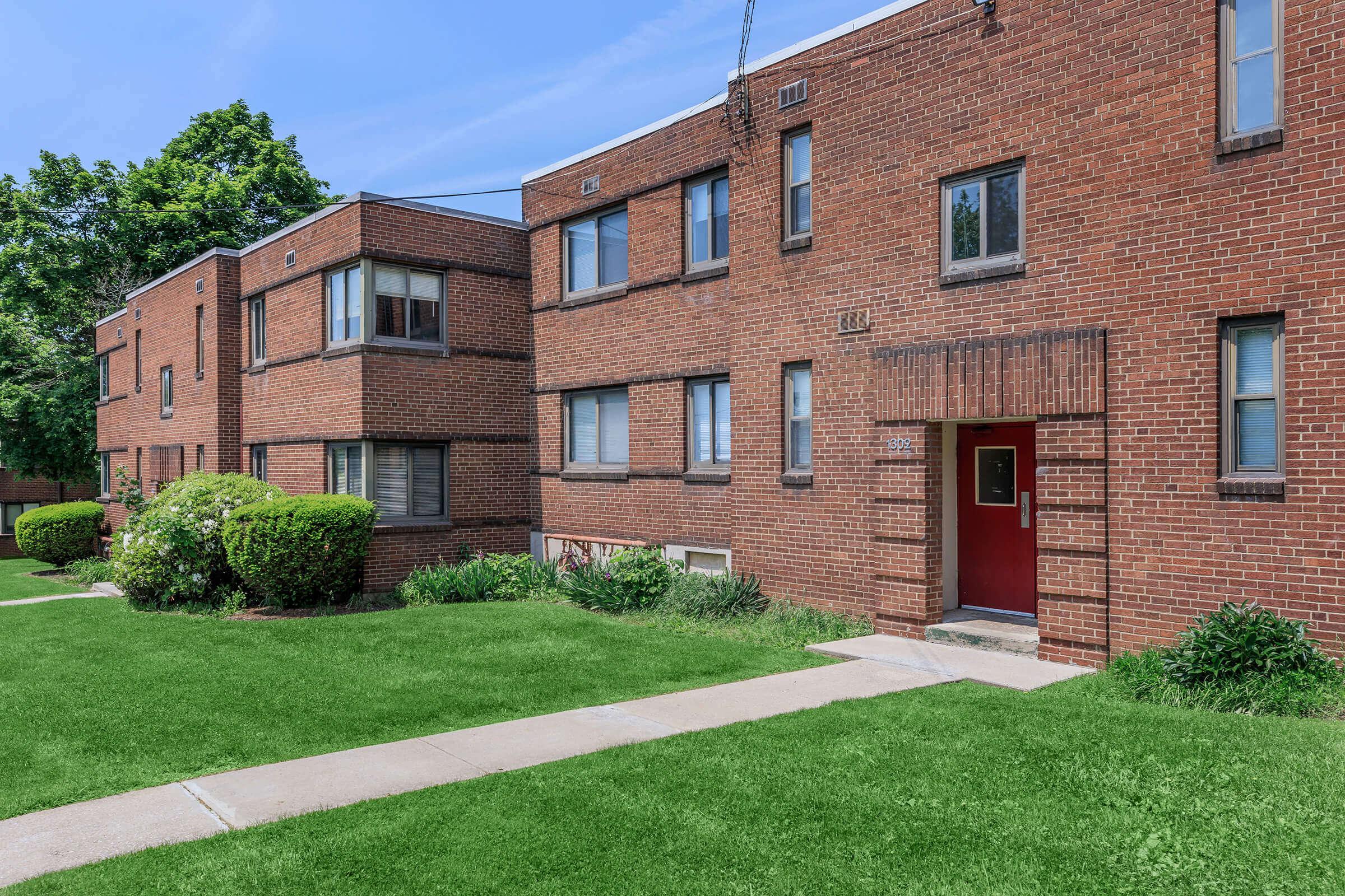 Brick apartment building with multiple windows and a red door, surrounded by green lawns and neatly trimmed bushes. The sky is clear and blue, and walkways lead to the entrance of the building.