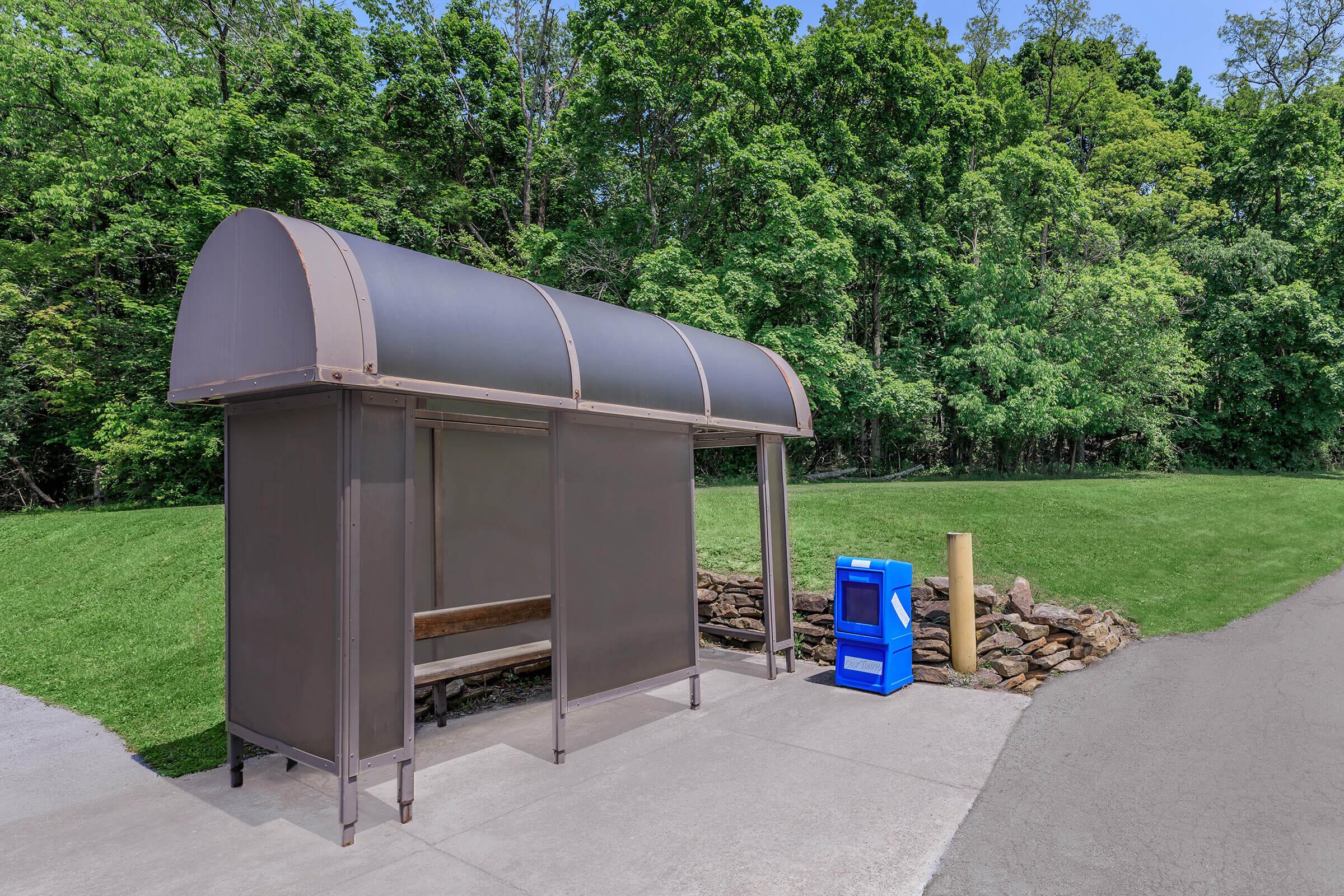 A bus stop shelter with a curved roof and a wooden bench sits alongside a paved path. Nearby, there is a blue newspaper stand and a grassy area bordered by trees in the background, creating a serene outdoor setting.
