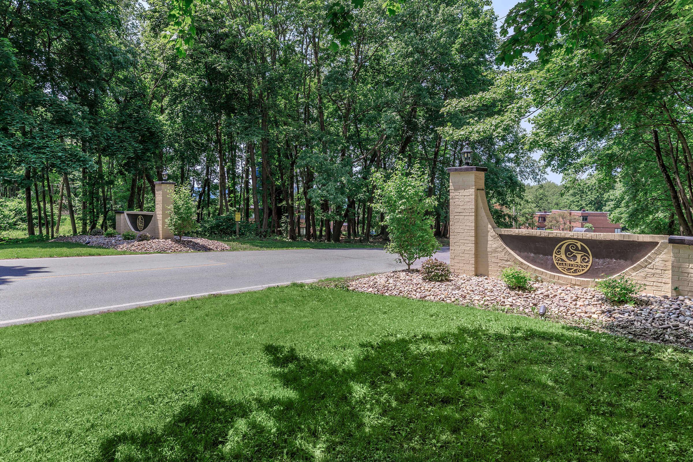 Entrance to a wooded area featuring a paved road flanked by decorative stone pillars. The pillars display an emblem and are surrounded by landscaped greenery, including shrubs and small rocks, under a clear sky.