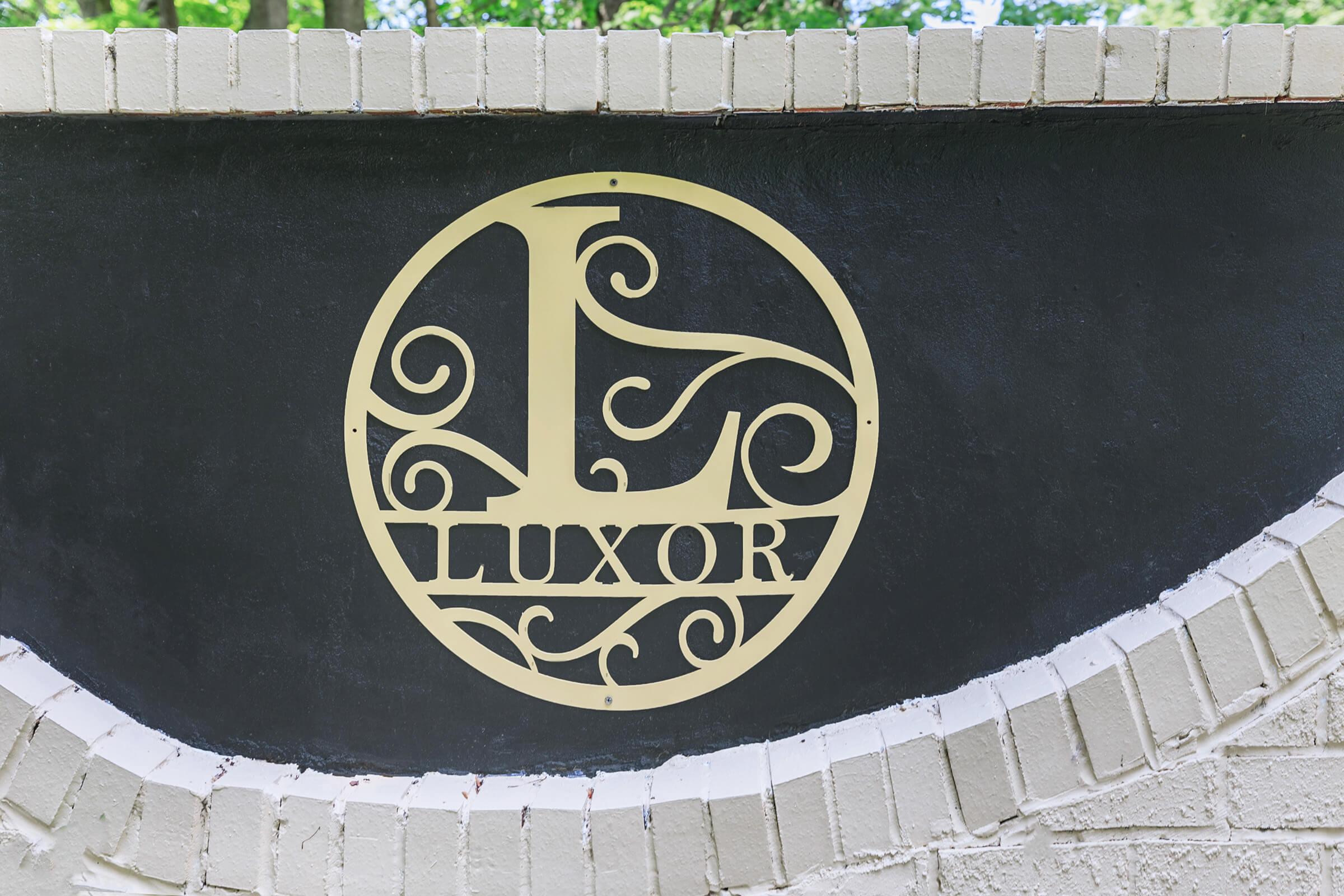 Sign featuring a decorative gold emblem with the letter "L" intertwined in a circular design, accompanied by the word "LUXOR" in an elegant font. The sign is mounted on a textured black surface surrounded by light-colored bricks. Glistening trees are visible in the background.