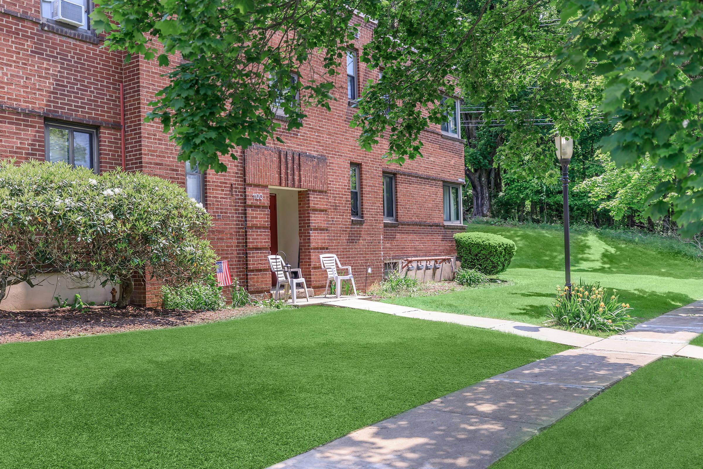 A well-maintained brick apartment building with green landscaping. Two white lawn chairs are positioned near the entrance, and a pathway leads to the front door. The scene features a lush green lawn and a few trees, creating a welcoming and serene atmosphere.