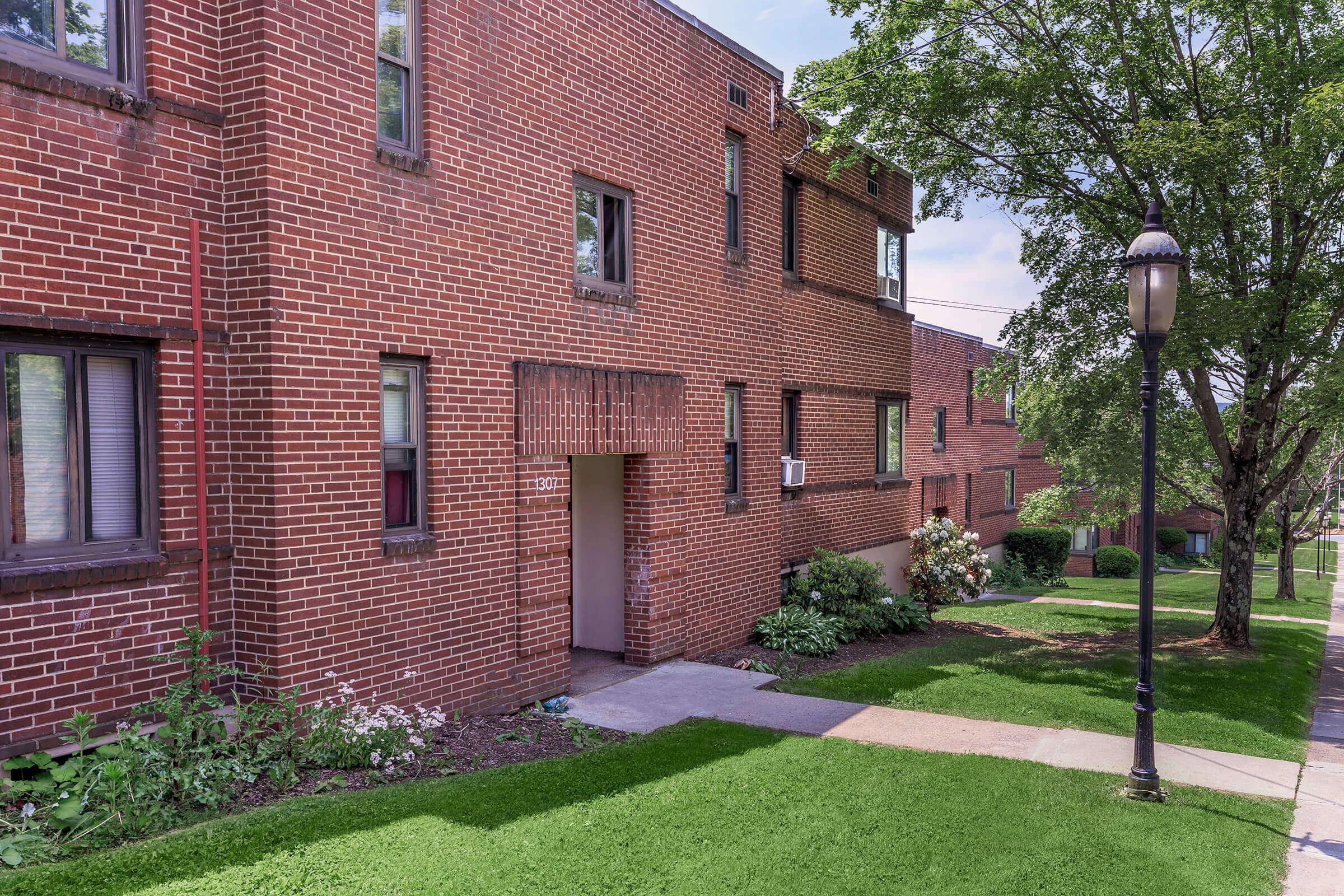 A brick apartment building with several windows and a doorway. Green grass and small bushes surround the entrance, and a lamp post stands near the sidewalk. Trees provide shade in the vicinity, creating a pleasant outdoor environment.