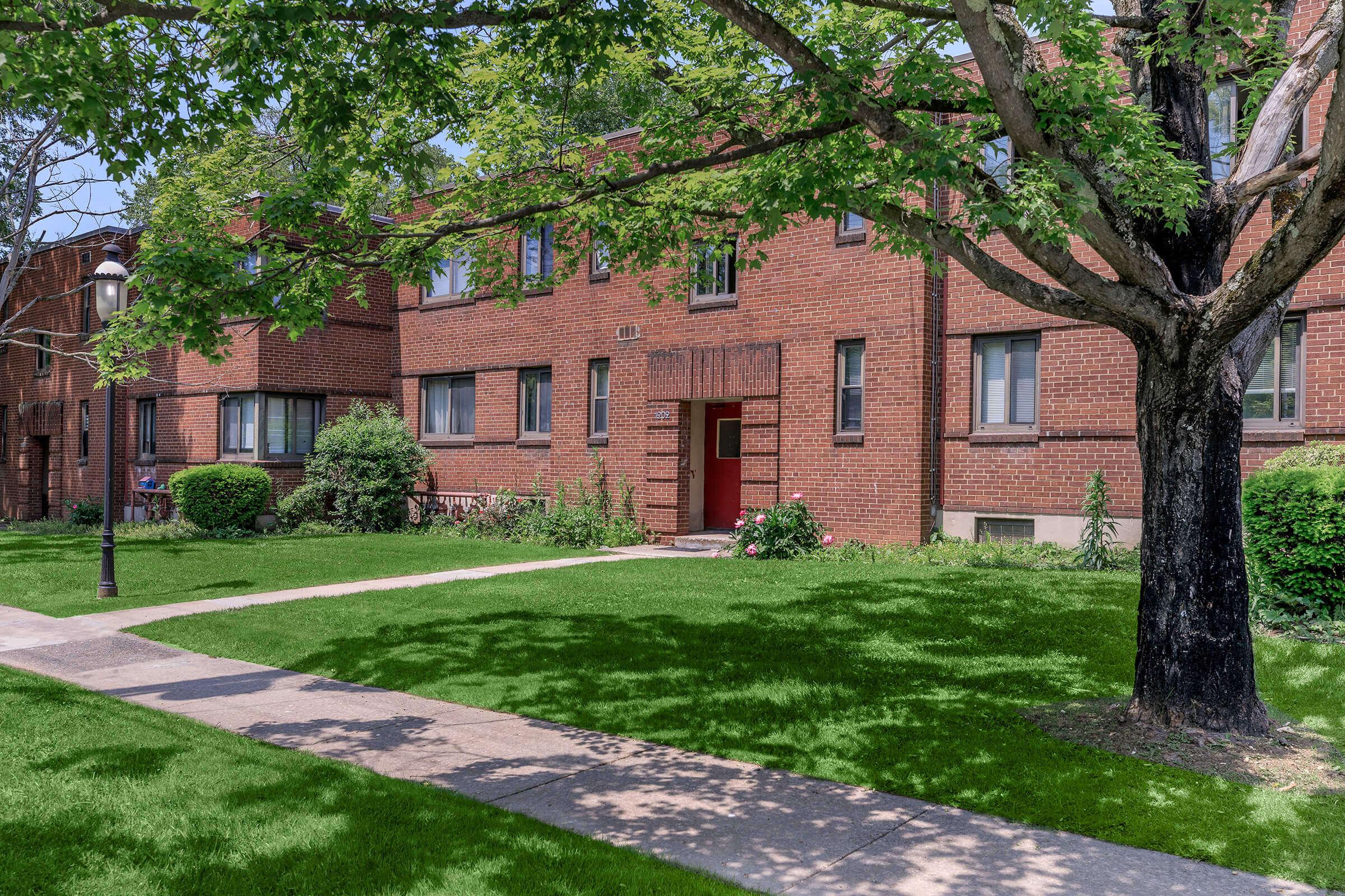 A brick apartment building surrounded by lush green grass and trees. A neatly paved sidewalk leads to the entrance, while shrubs and landscaping add to the exterior charm. The setting is bright and inviting, typical of a residential area.