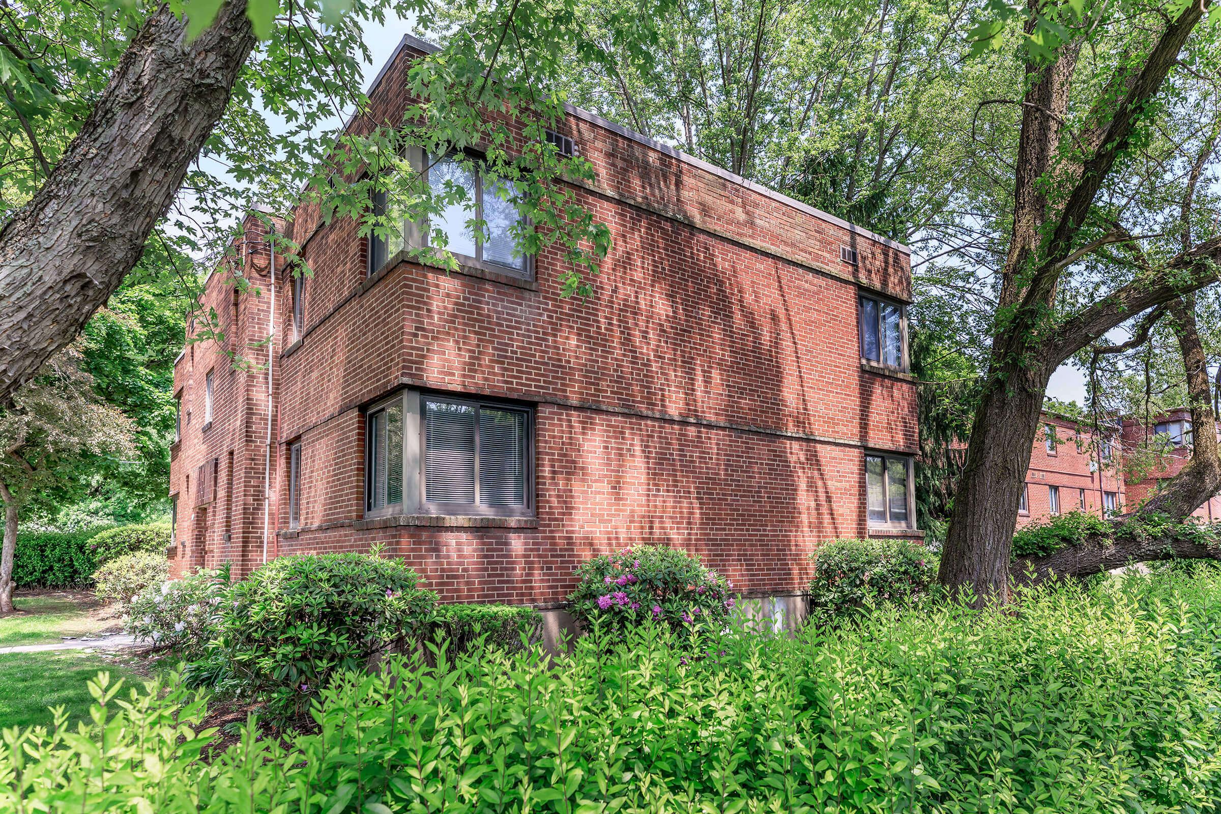 A brick apartment building surrounded by lush greenery and trees. The building features large windows and a blend of shaded and sunlit areas, with well-maintained shrubs and flowers in the foreground, creating a welcoming residential environment.