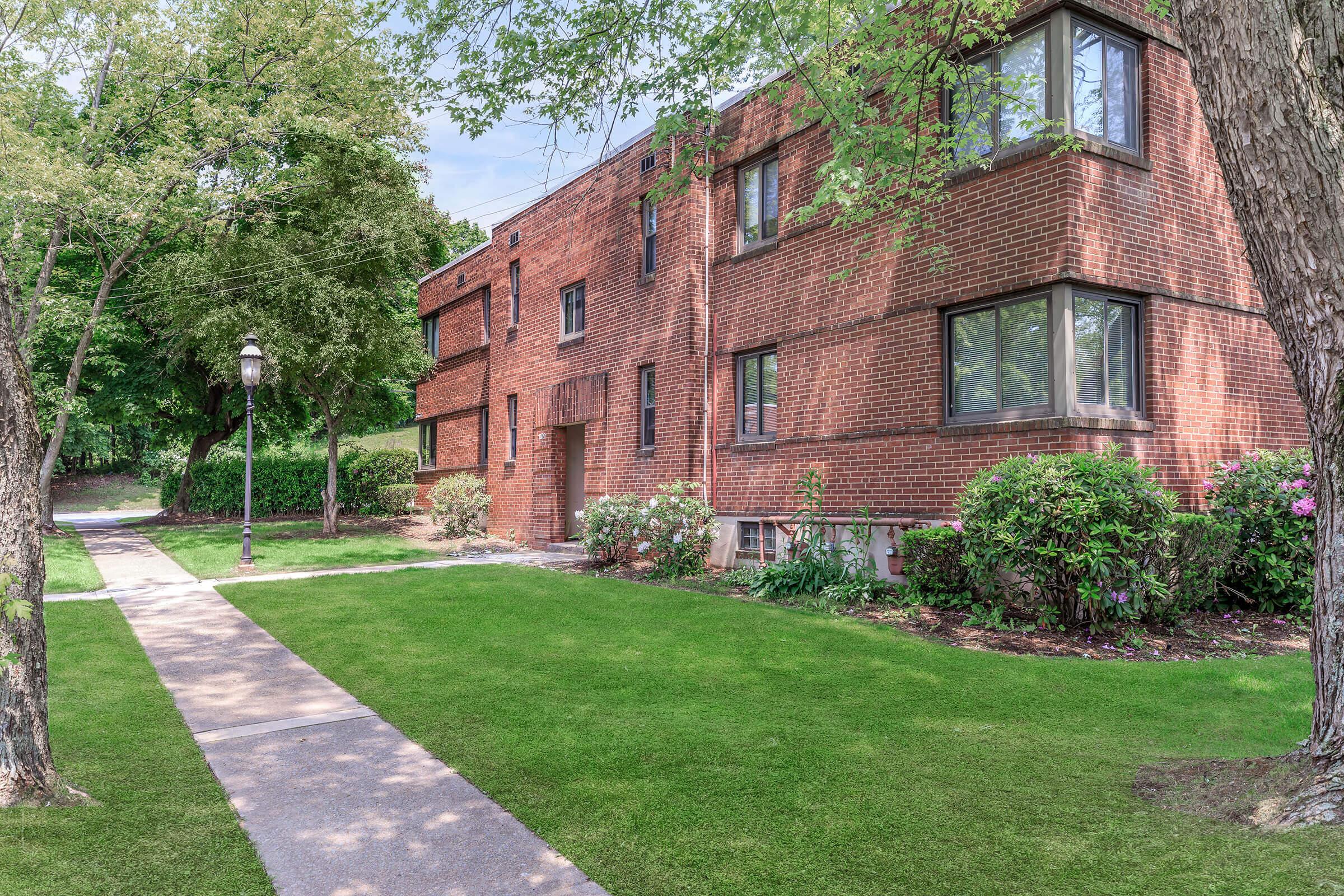 A residential brick building surrounded by greenery, with a well-maintained lawn and flowerbed. A concrete walkway leads to the entrance, and there are trees providing shade in the surrounding area. The scene is bright and inviting, suggesting a pleasant outdoor environment.