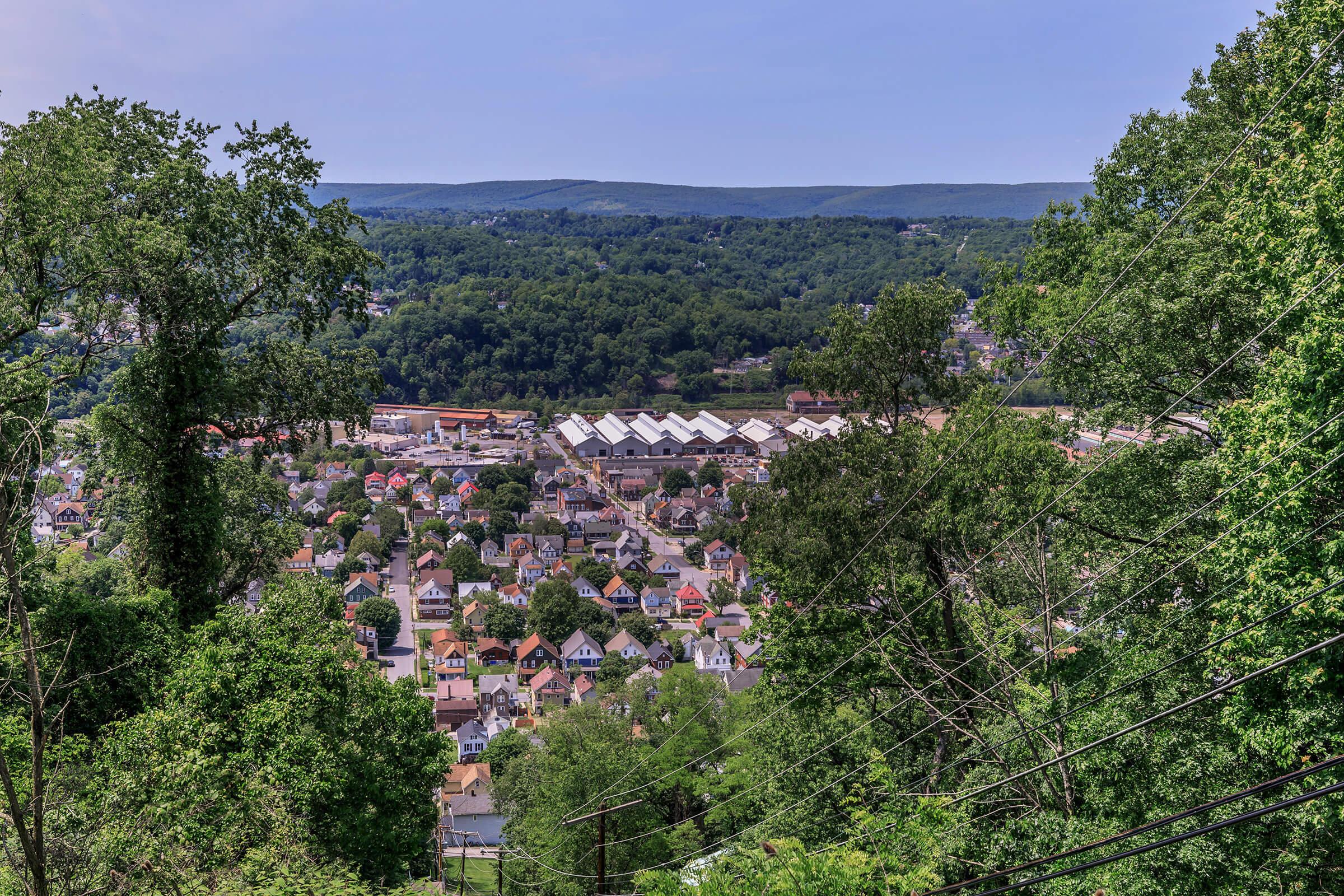 A panoramic view of a small town nestled in a valley surrounded by dense green hills. The foreground shows a variety of residential buildings with colorful rooftops, while in the background, the landscape features rolling hills under a clear blue sky. Power lines run diagonally across the image.