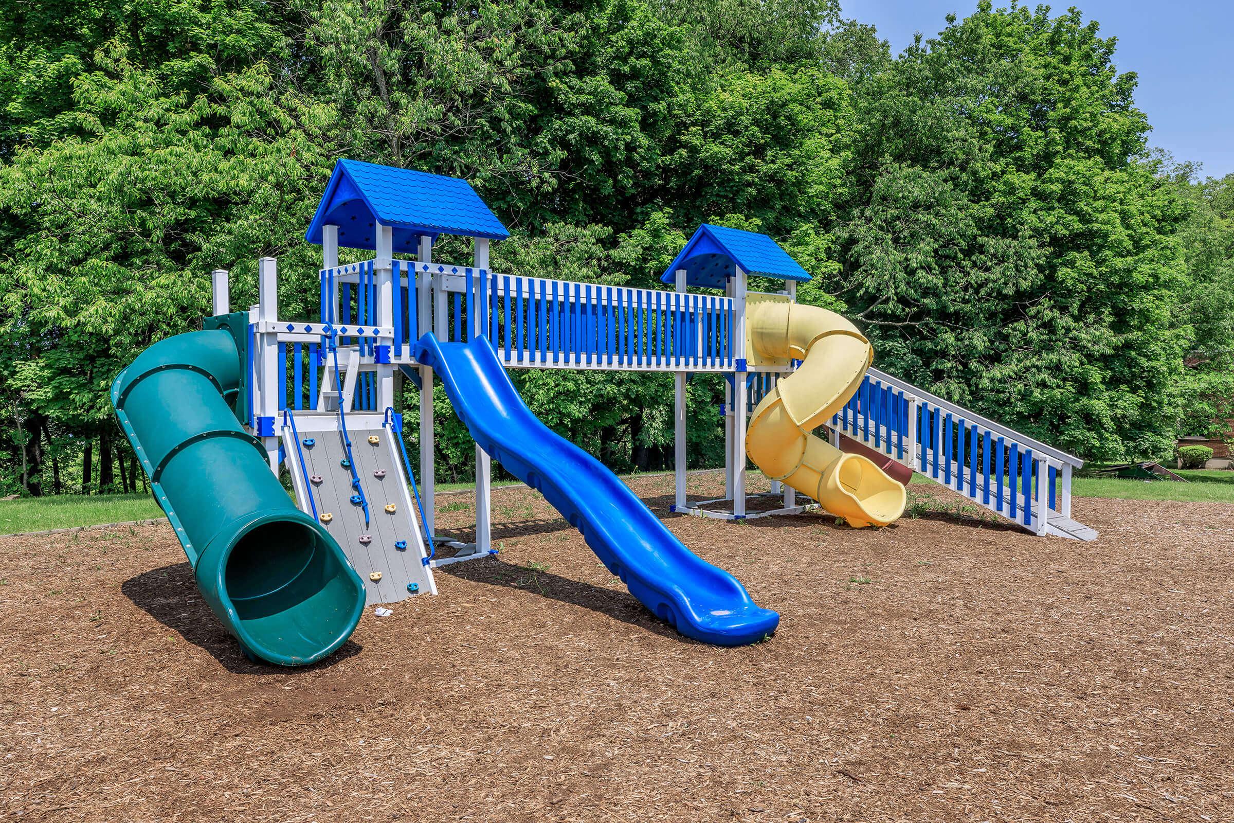 A colorful playground structure featuring blue, yellow, and green slides, climbing walls, and a green tube slide. The playground is surrounded by a mulch surface and lush green trees in the background, under a clear blue sky. Ideal for children's outdoor play and recreation.