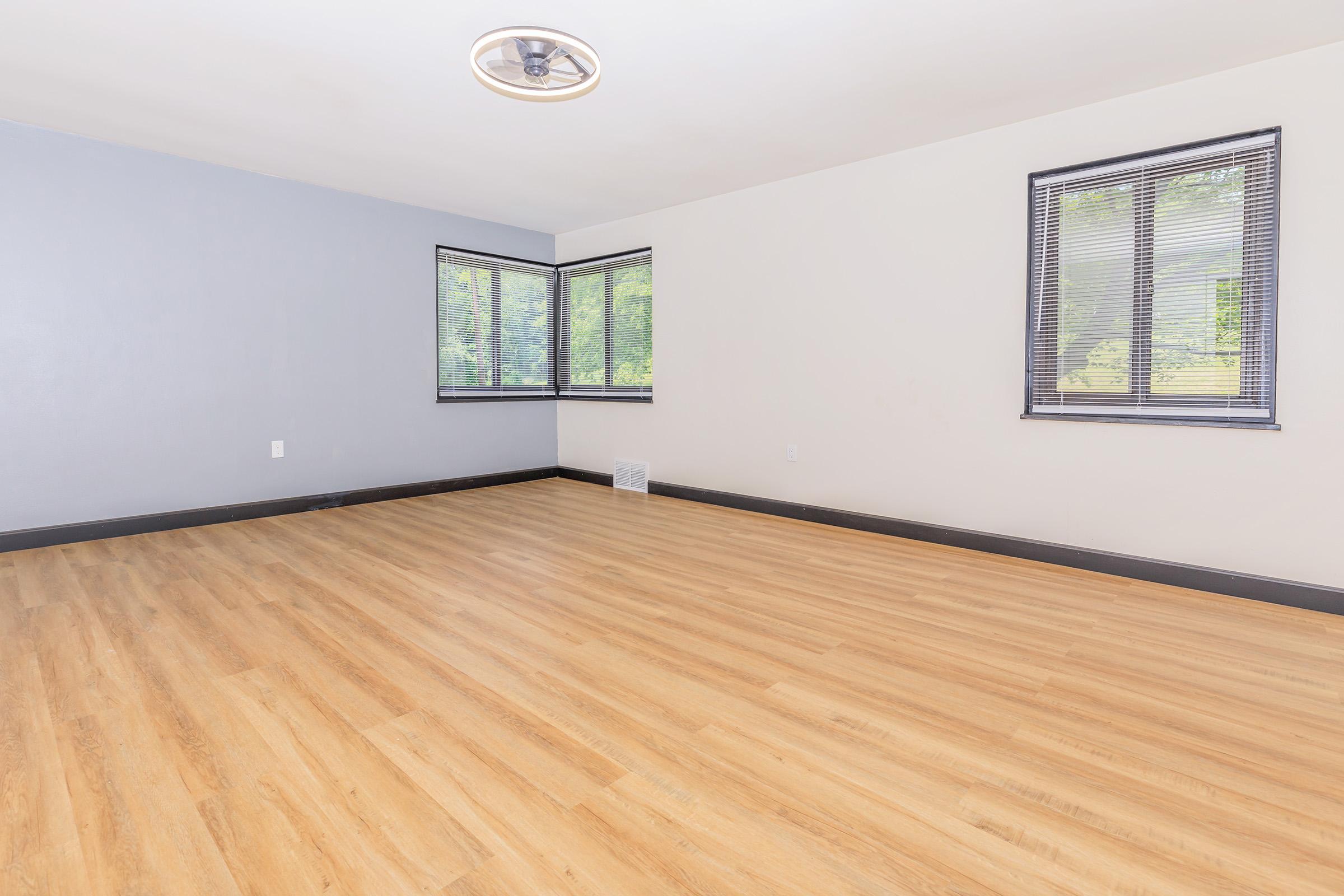 A spacious, empty room featuring light wood flooring, gray walls, and large windows with blinds. The room has minimal decoration and is brightly lit, creating an open and airy atmosphere.