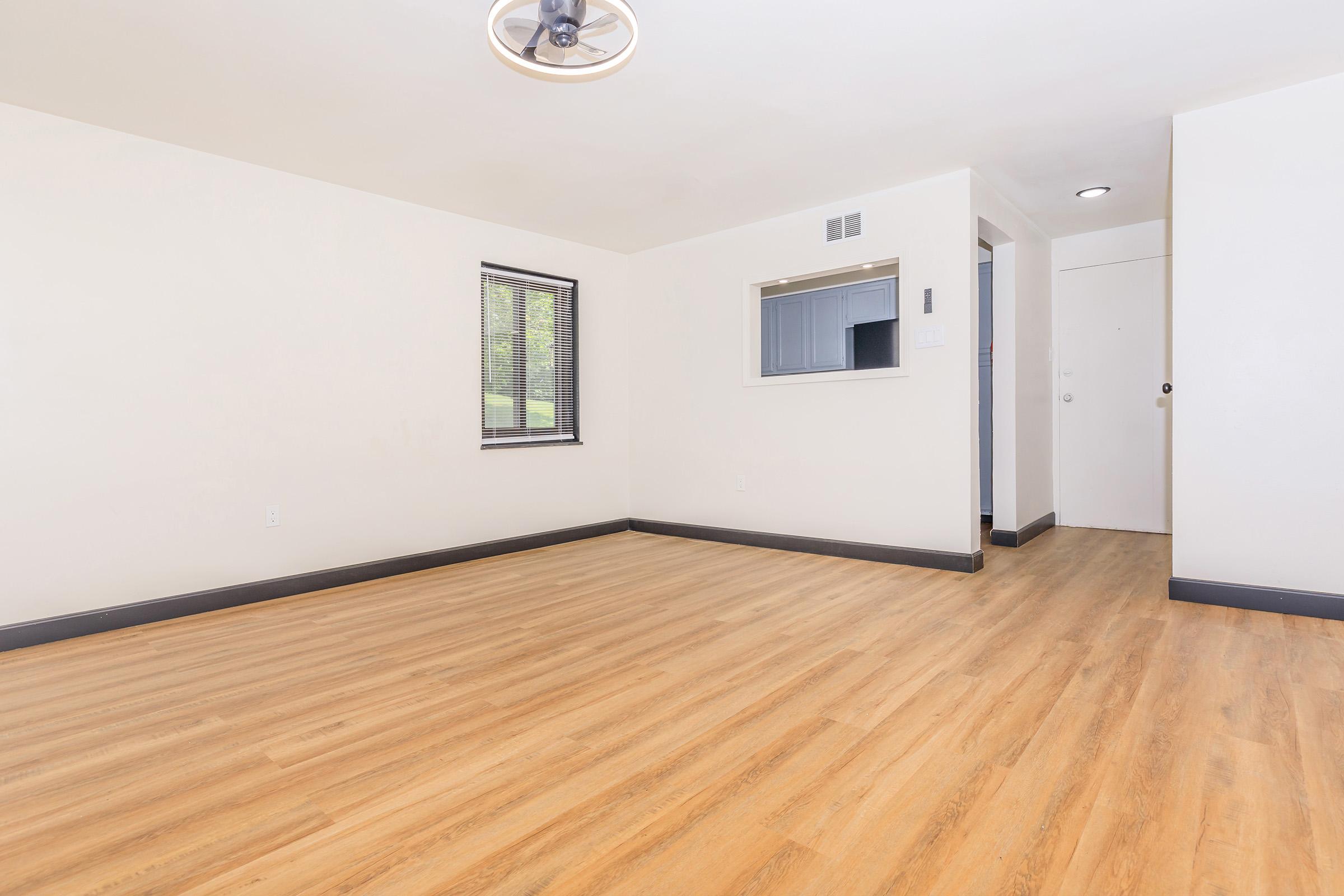 A spacious, empty room with light-colored walls and a wooden floor. A ceiling fan is visible above, and there is a window on the left side letting in natural light. A simple door is seen on the right, and a small opening with a counter can be seen in the background, hinting at a kitchen or service area.
