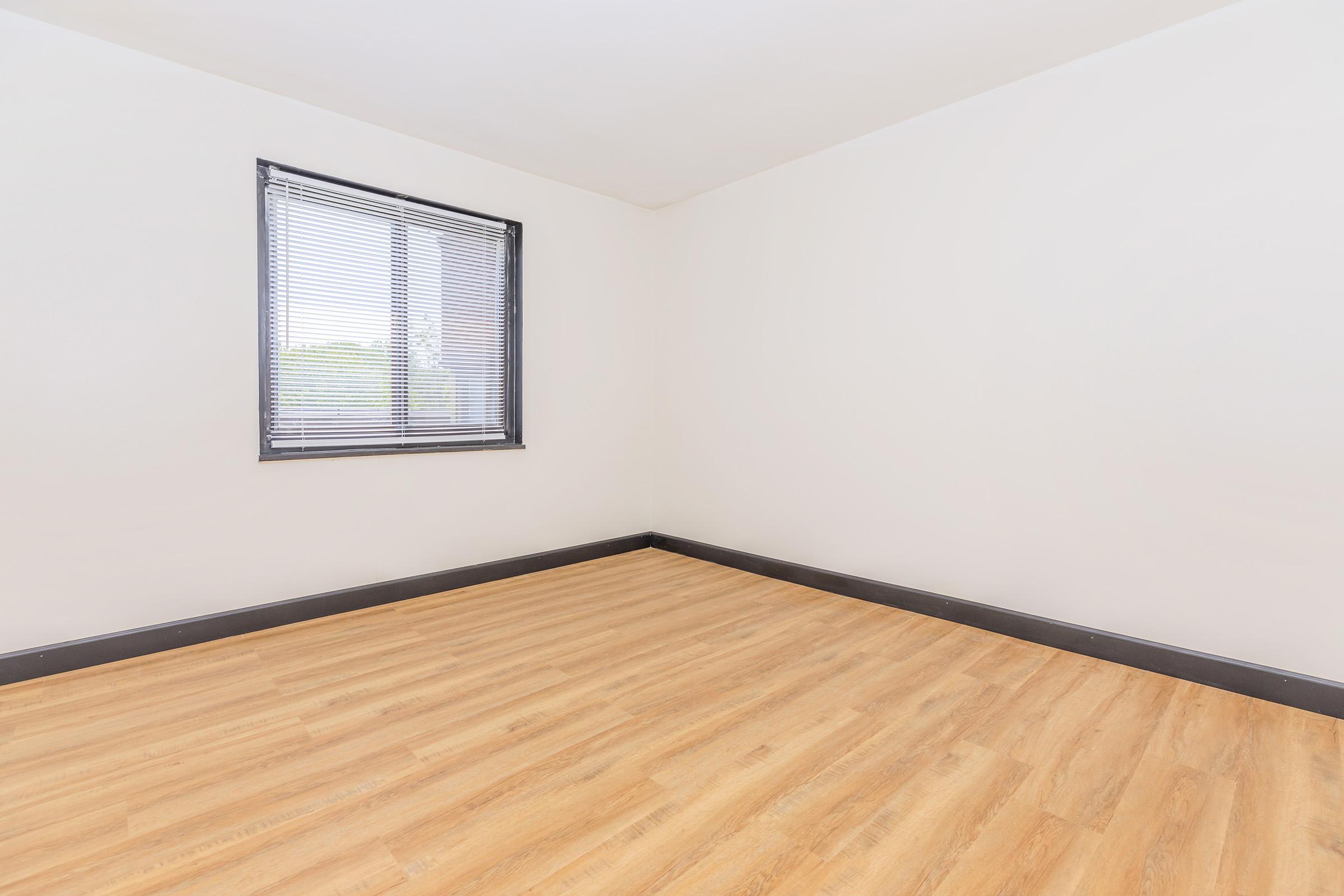 Empty room with light-colored walls and a medium-brown wooden floor. A window with horizontal blinds is on one wall, allowing natural light to enter. The space has no furniture or decor, creating a minimalist and open feel.