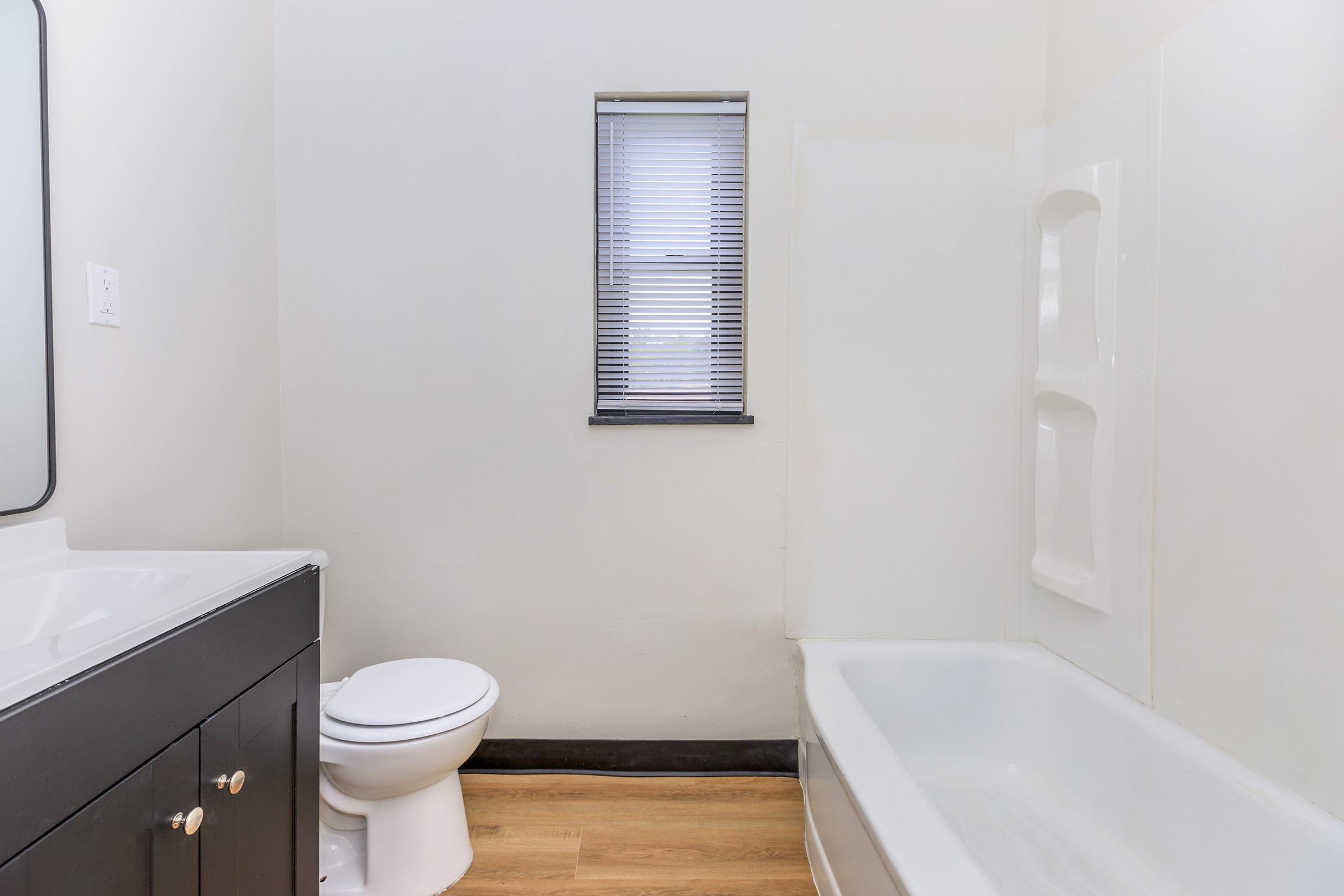 A clean and modern bathroom featuring a white bathtub, a dark-colored vanity with two cabinet doors, a toilet, and a window with blinds letting in natural light. The walls are painted light, creating an airy feel, and the floor is a warm-toned wood laminate.
