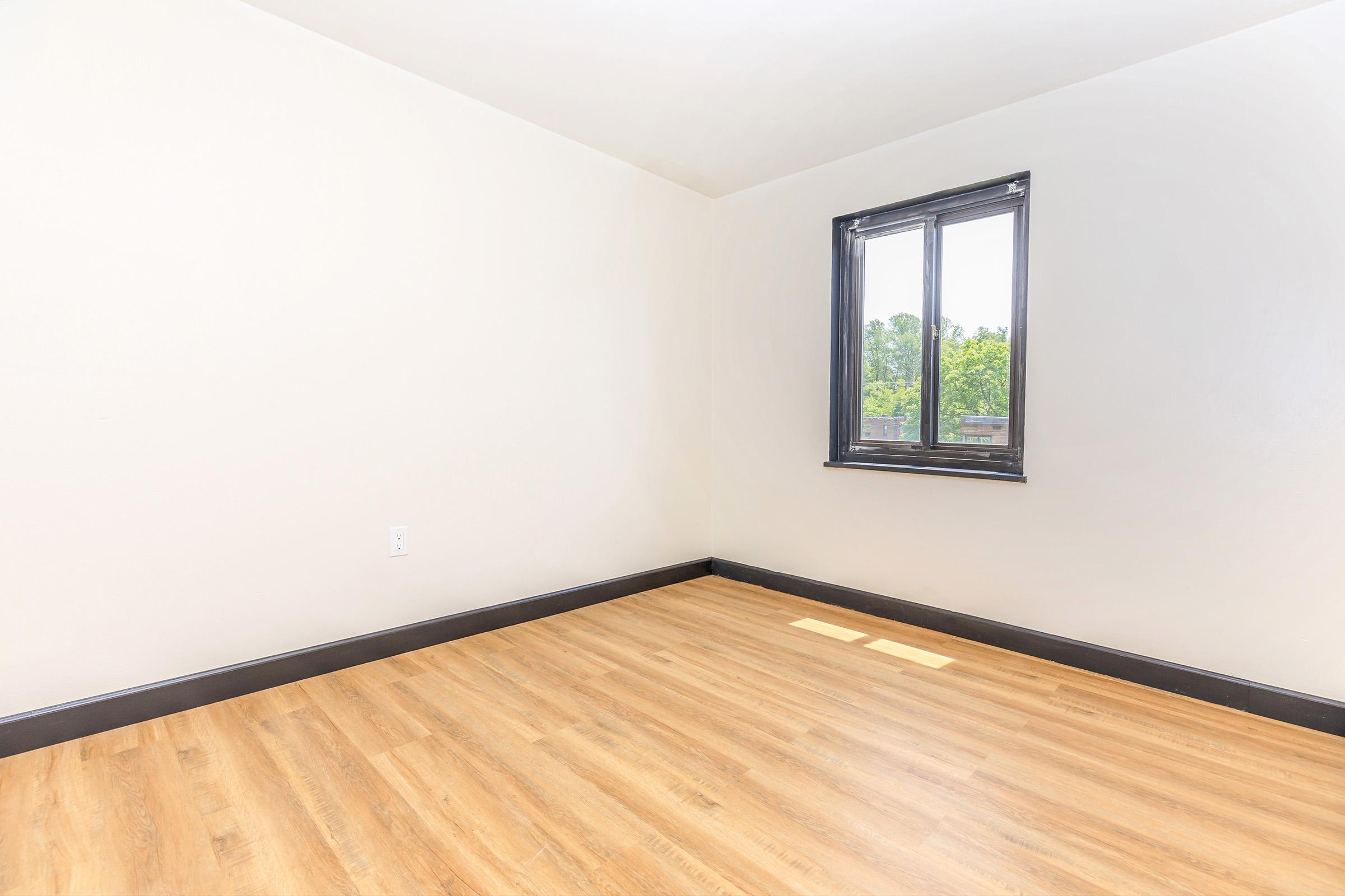 Empty room featuring a light wooden floor, cream-colored walls, and a dark trim around the window. A single window allows natural light to enter, providing a view of greenery outside. The space is minimalistic and ready for decoration or furnishing.