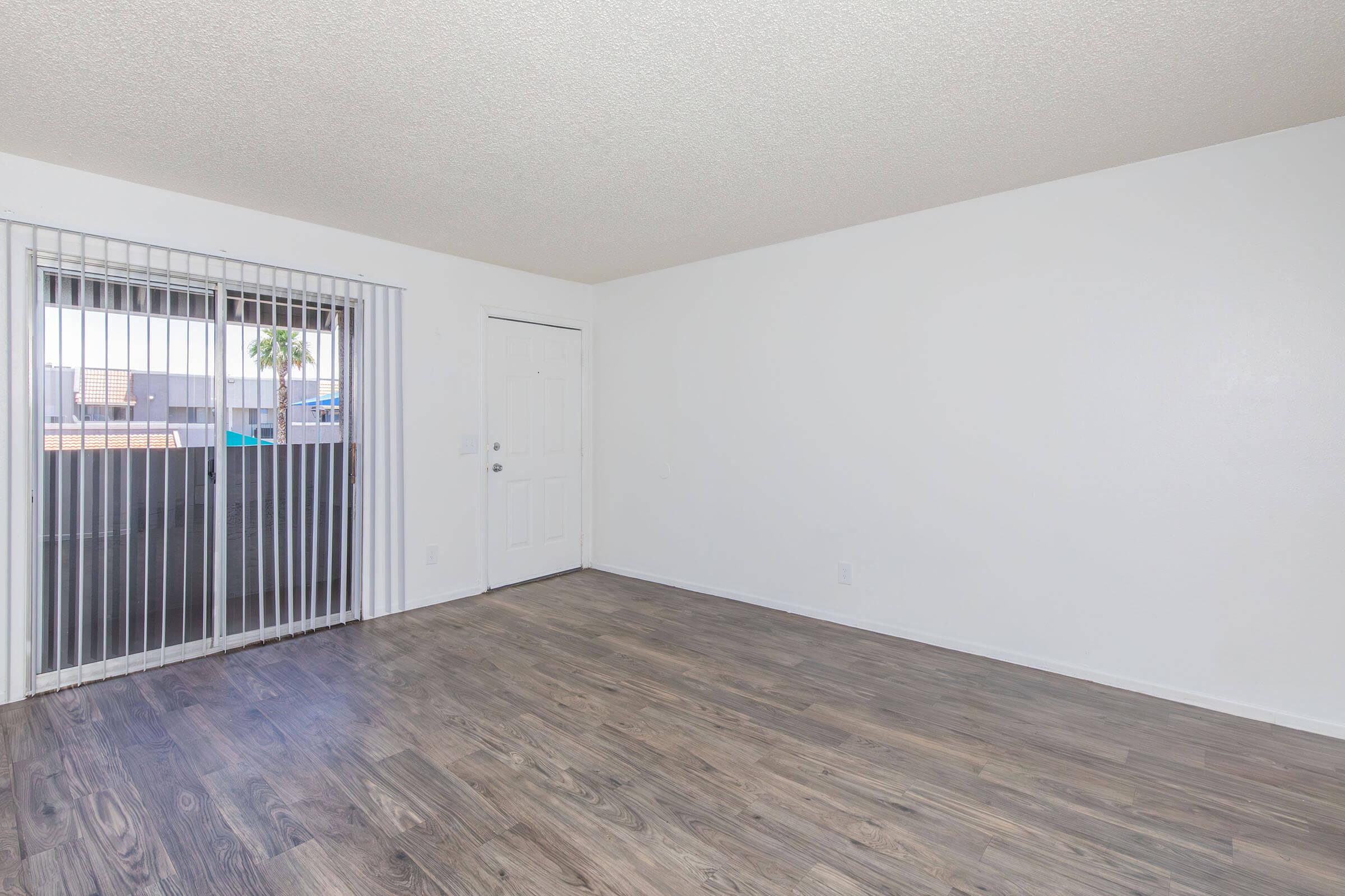 An empty room with light-colored walls and a wooden floor. A sliding glass door with vertical blinds leads to an outdoor area, and a white door is visible on the right. The room has minimal furnishings, creating an open and spacious feel. Natural light enters through the sliding door.
