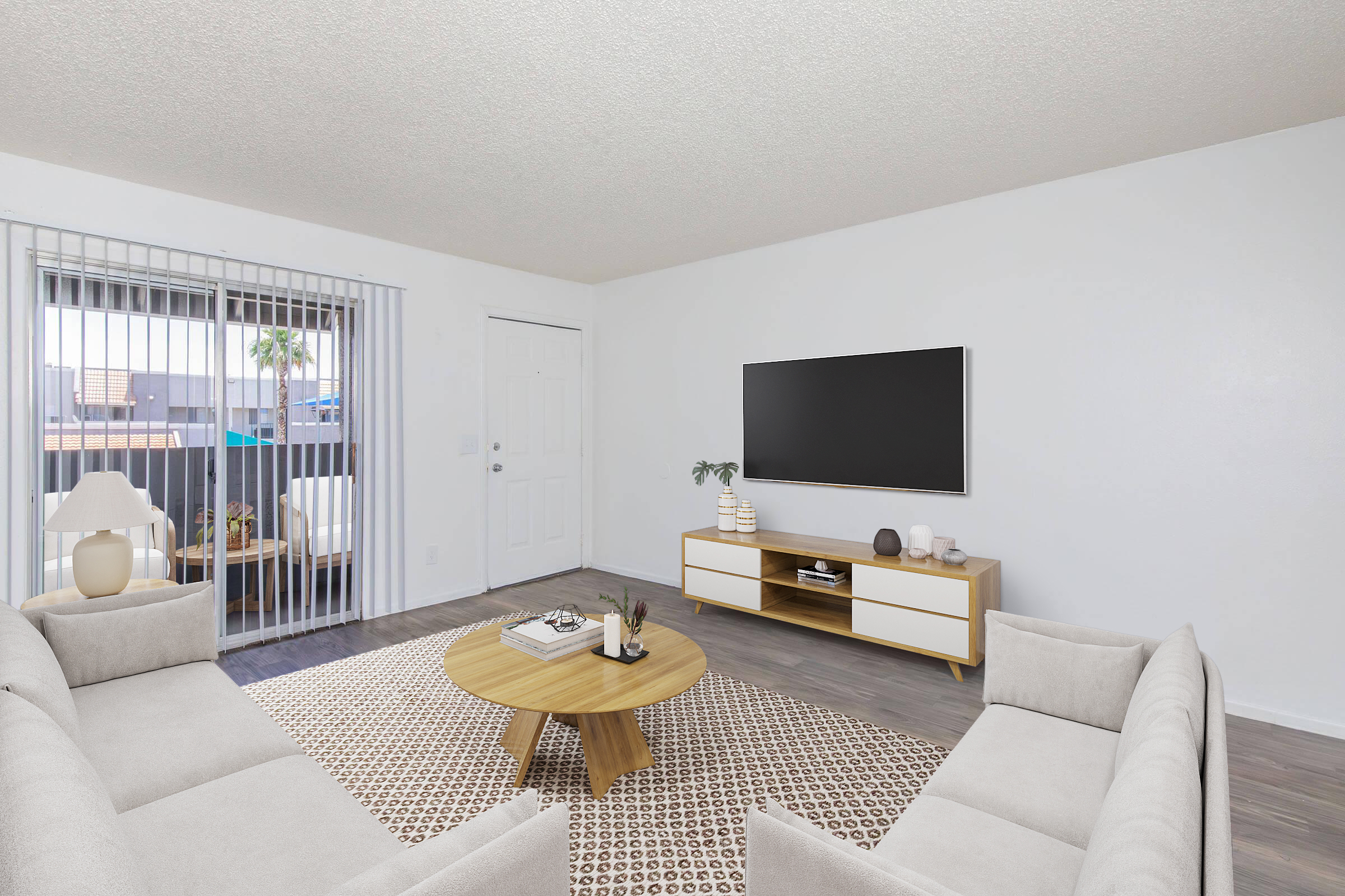 A modern living room featuring two light gray sofas facing a round wooden coffee table. A television is mounted on a light wood media console against a white wall. Large sliding doors lead to a balcony with outdoor seating. Natural light brightens the space, complemented by a patterned area rug.