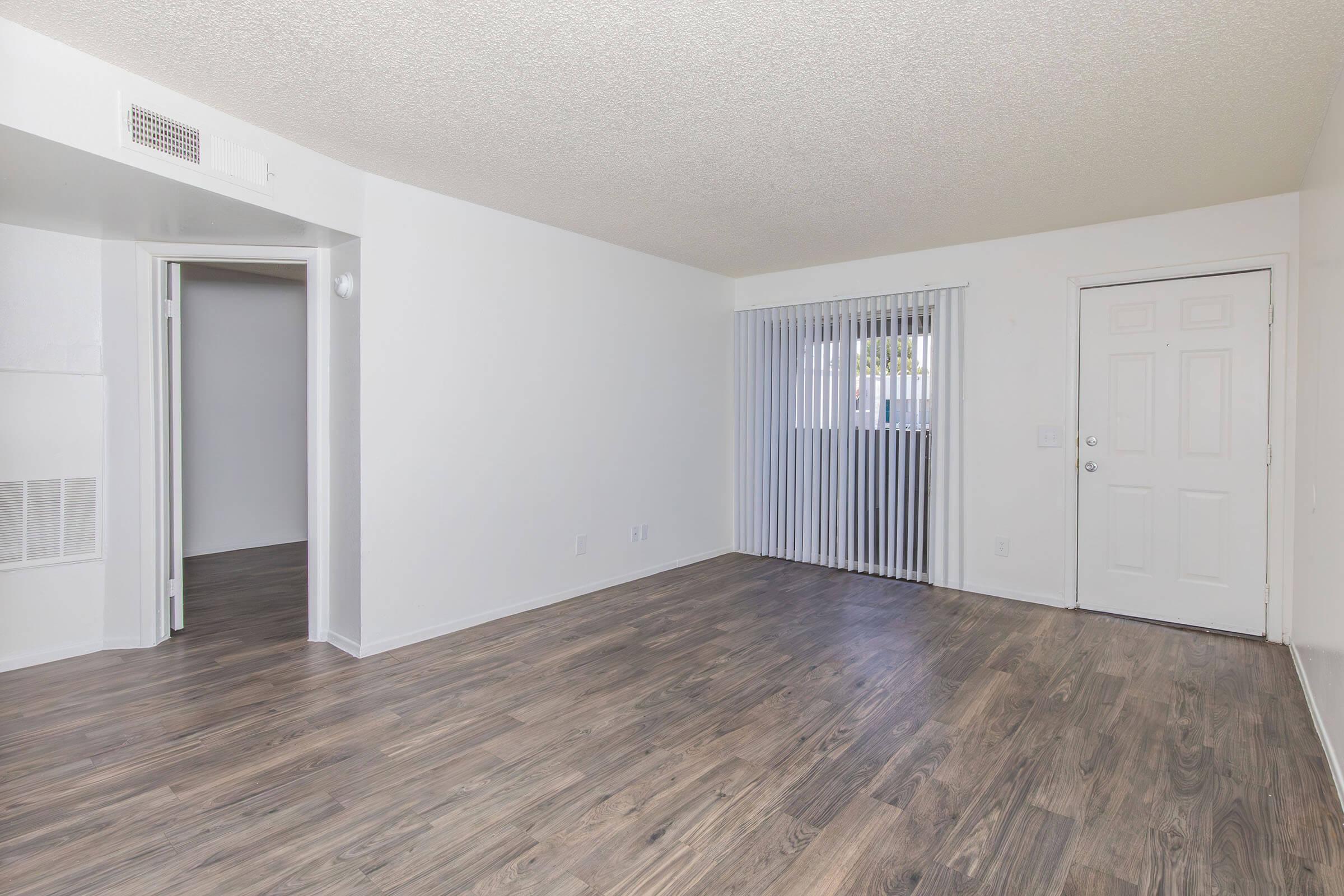 Empty living room featuring light-colored walls, a textured ceiling, and laminate flooring. A door on the right leads outside, while vertical blinds cover a window on the left. A small hallway entrance is visible, leading to another room, creating a spacious and bright atmosphere.