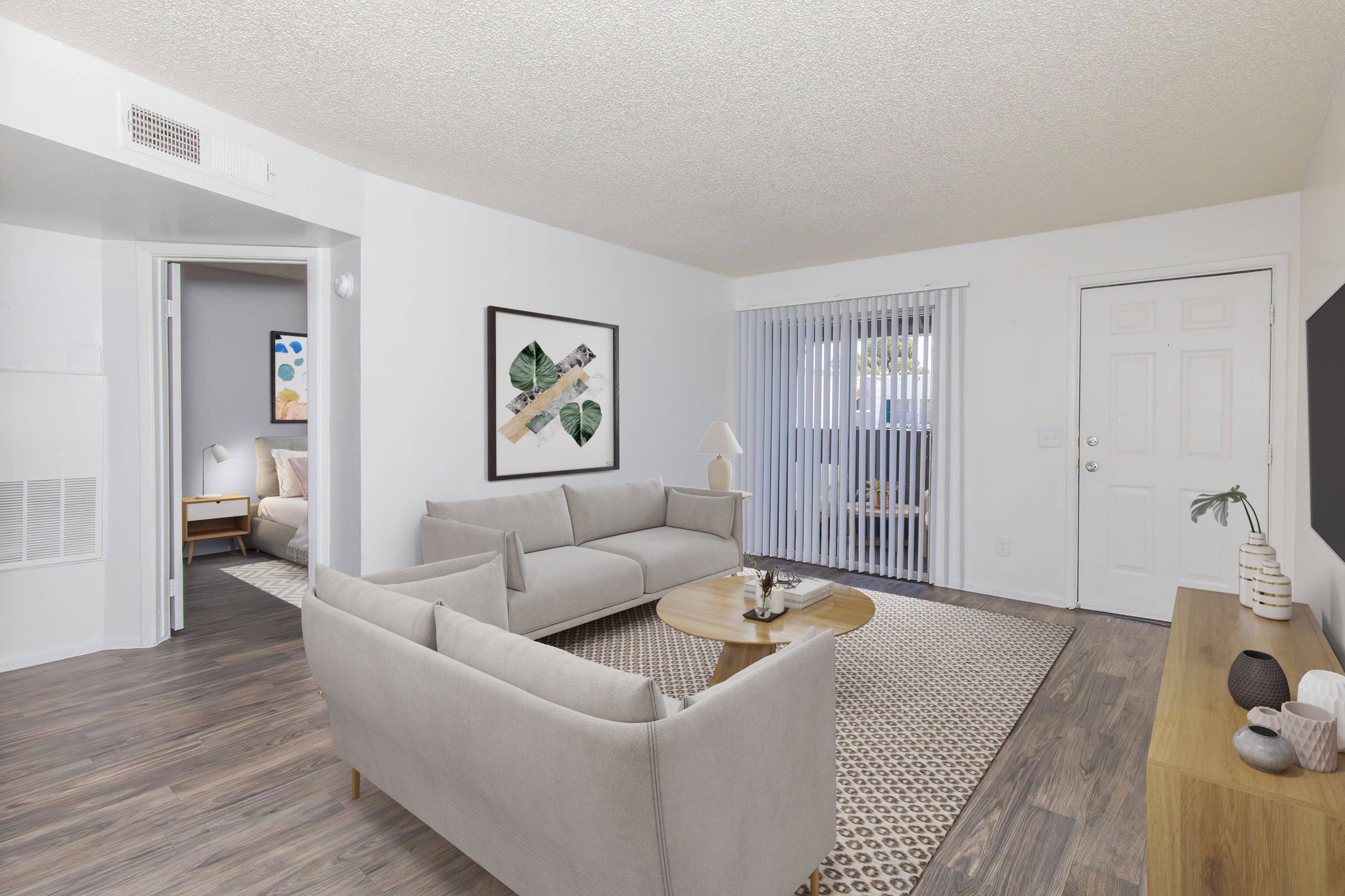 A bright, airy living room featuring a light gray sofa, a round wooden coffee table, and a flat-screen TV on a wooden console. There are potted plants and artwork on the walls. Large windows with sheer curtains allow natural light, and a doorway leads to a bedroom area in the background.