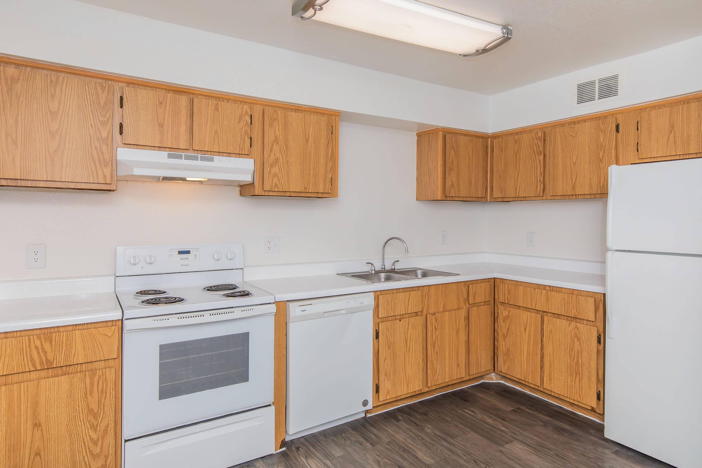 Modern kitchen featuring wooden cabinets, a white stove with an oven, a dishwasher, and a refrigerator. A double sink is positioned under the window, with countertops designed for efficiency. The light fixtures provide bright illumination, enhancing the clean and organized space.