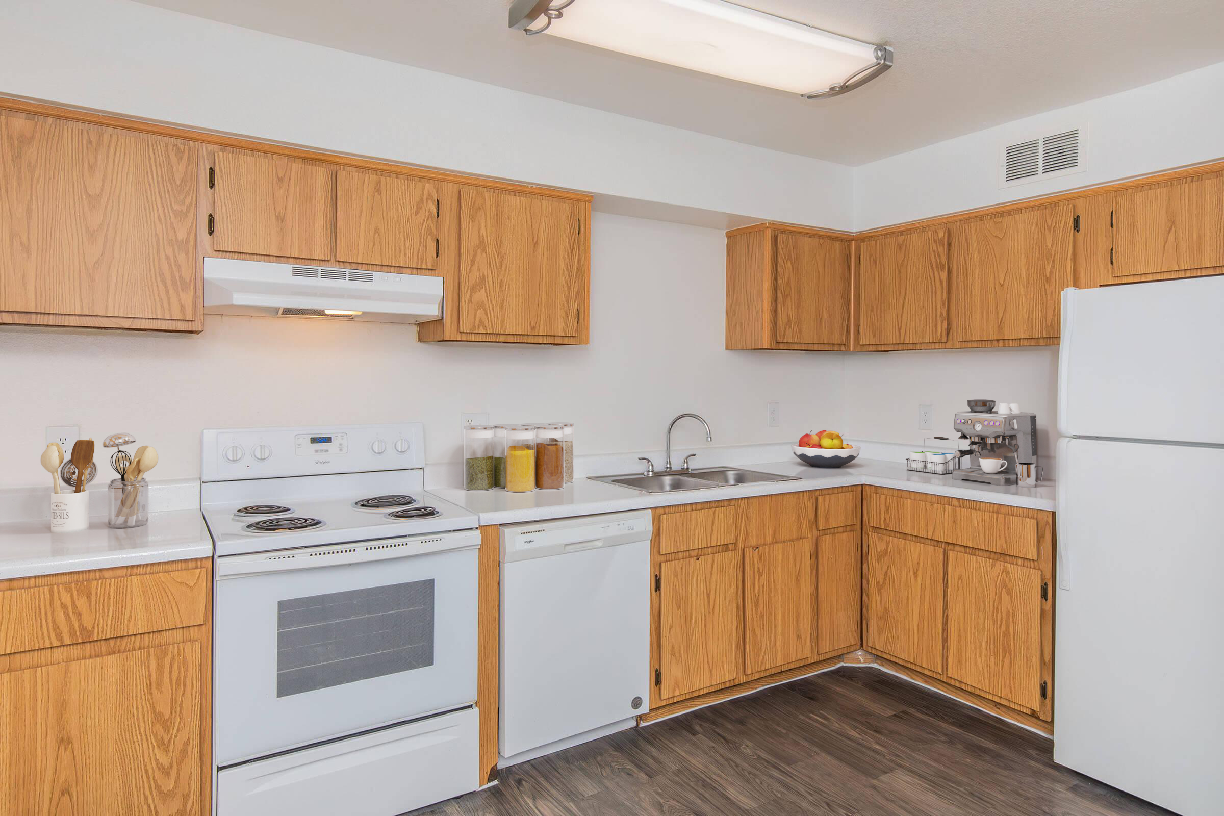 Modern kitchen with wooden cabinets, a white countertop, and stainless steel appliances. Features include a stove, microwave, dishwasher, and refrigerator, alongside a sink and a bowl of fruit on the counter. Various jars of ingredients are neatly arranged on the countertop.