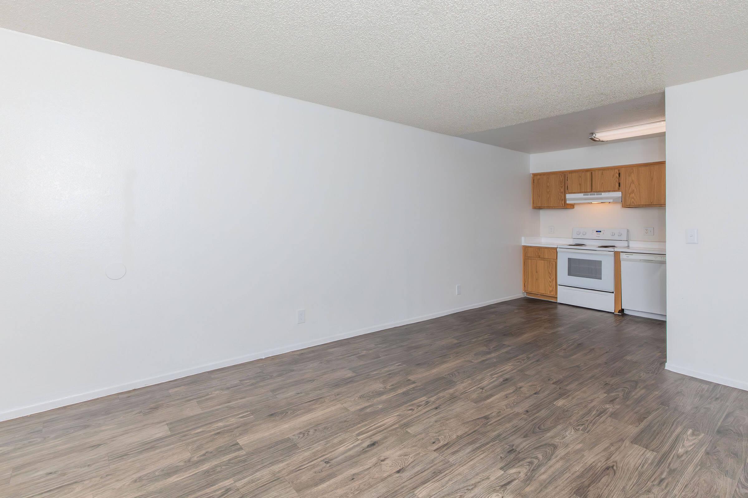 A spacious, empty room with light-colored walls and a wooden floor. In the background, there's a small kitchen area featuring wooden cabinets, a white stove, and a refrigerator. The area is well-lit with overhead lighting, creating a bright, open atmosphere.
