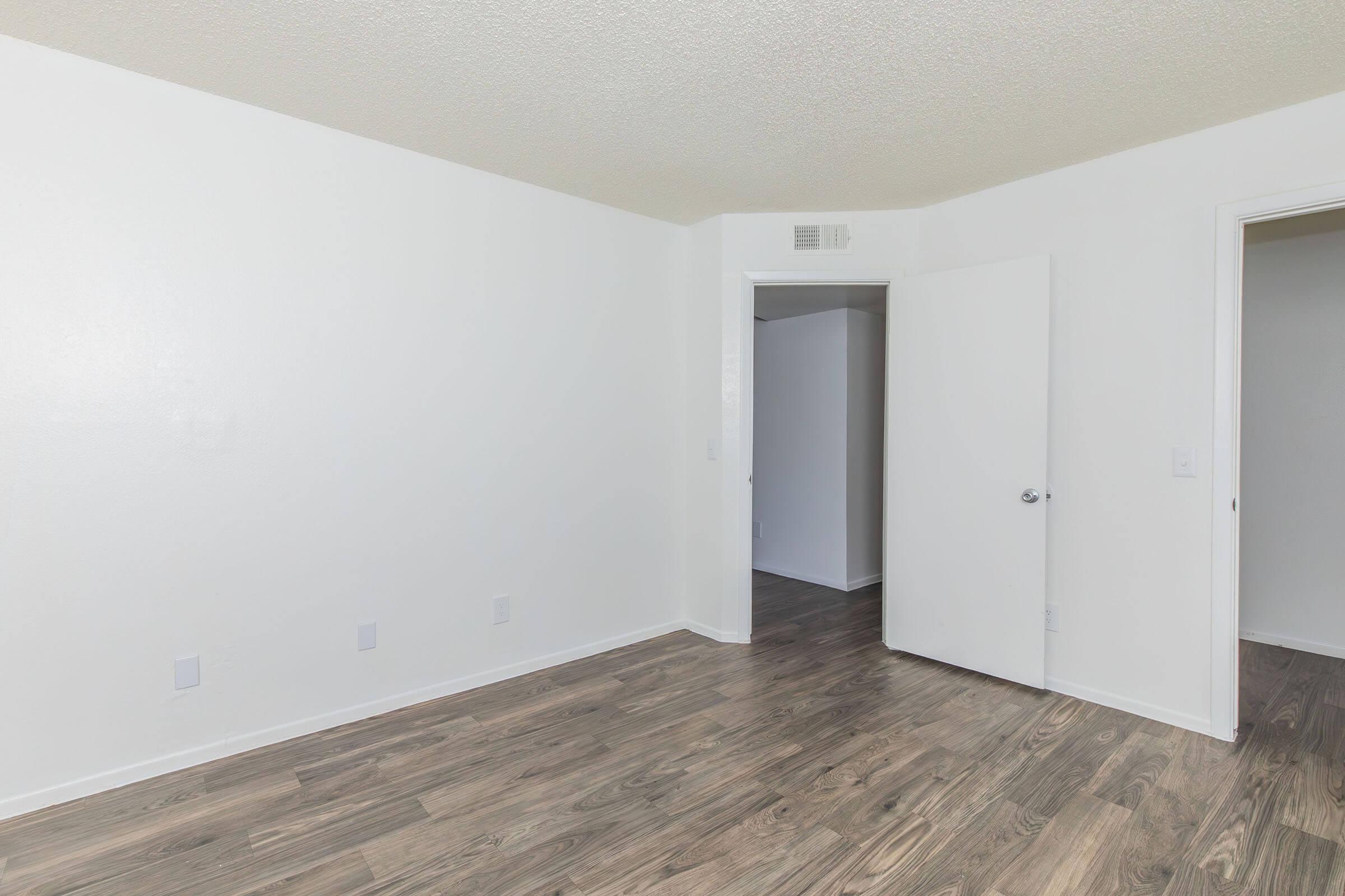 A partially empty room with light-colored walls and a textured ceiling. The floor features laminate wood, and there is a white door on the right side leading to another space. The room appears well-lit and spacious, with a minimalist design and no furniture.