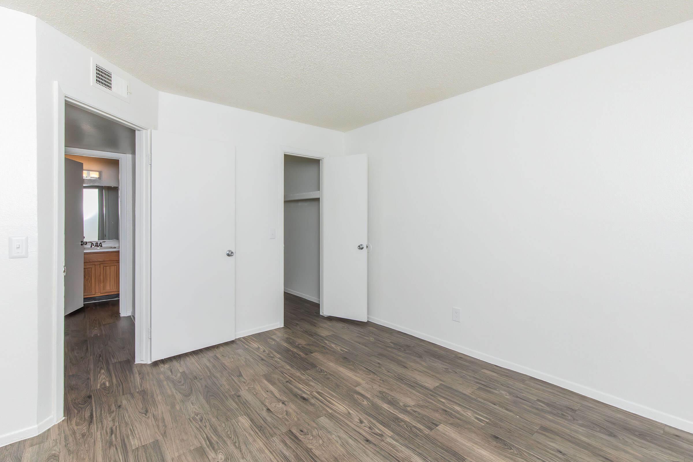 A spacious, empty room with light-colored walls and a textured ceiling. The floor is covered with hardwood-like laminate. There are two open closet doors on the left, and a doorway leads to a bathroom visible in the background. Natural light is coming from a window, creating a bright atmosphere.