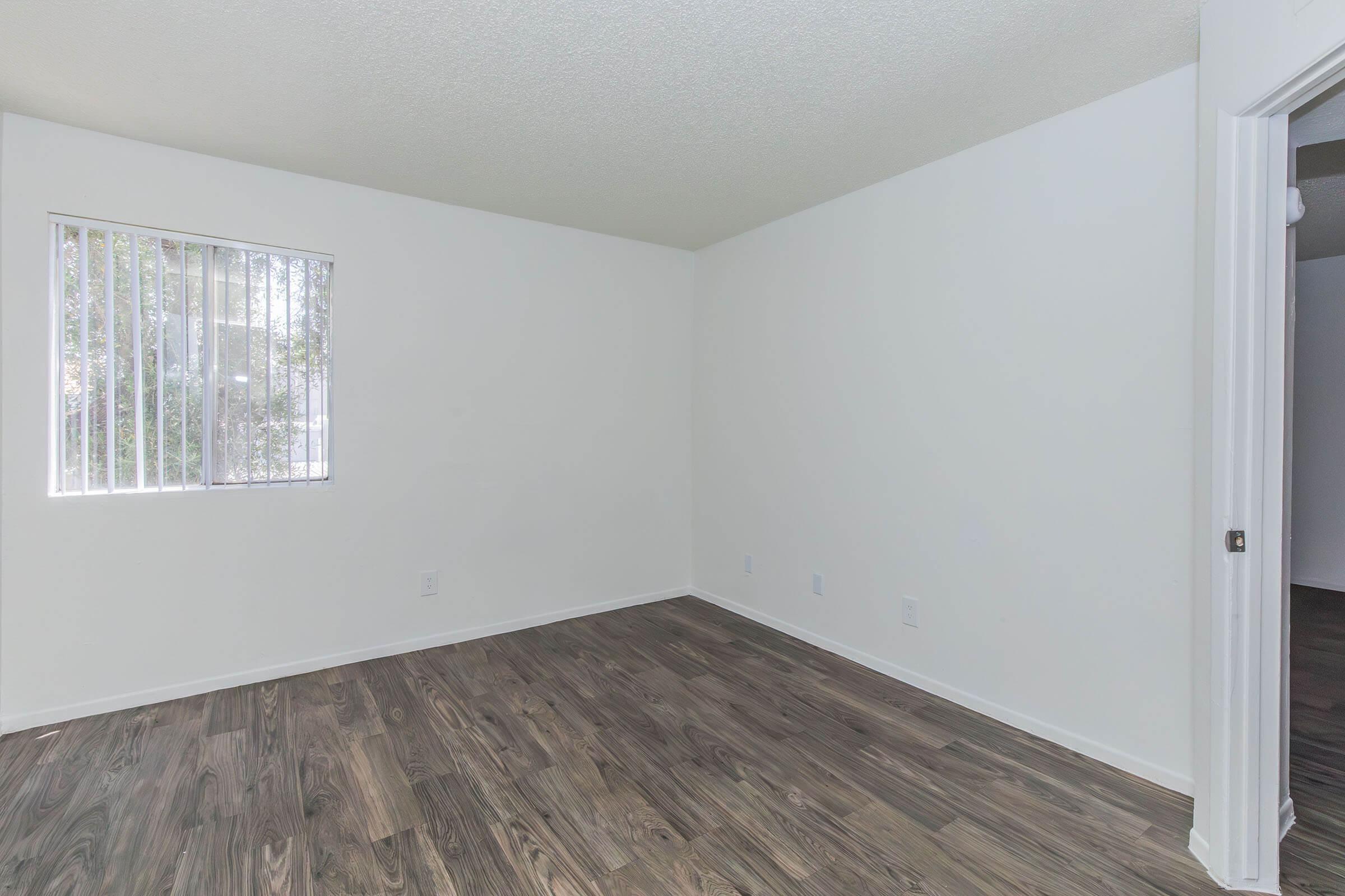 Empty room with light-colored walls and a window featuring vertical blinds. The floor is dark wood laminate, and there are no furnishings or decorations. The overall atmosphere is bright and clean, suitable for various purposes.