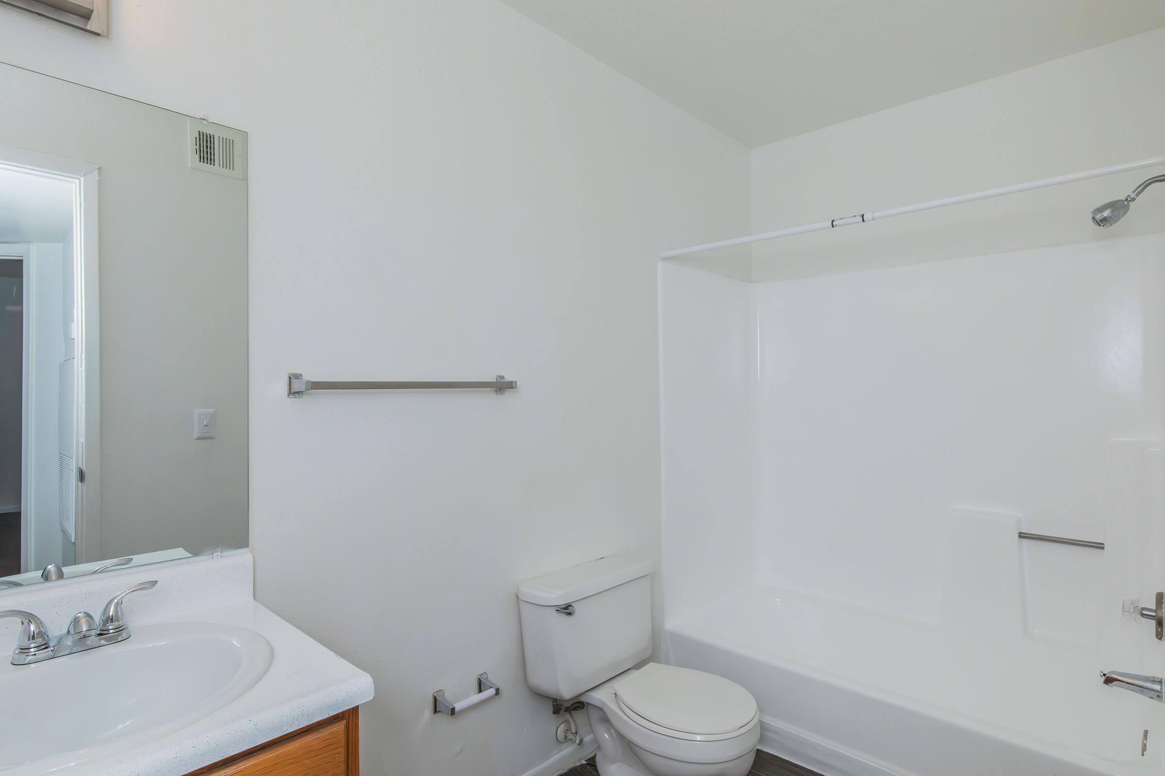 A clean and simple bathroom featuring a white bathtub with a shower, a toilet, and a sink with two faucets. The walls are painted white, and there's a mirror above the sink. A towel bar is mounted on the wall, and the floor appears to be a dark-colored material.