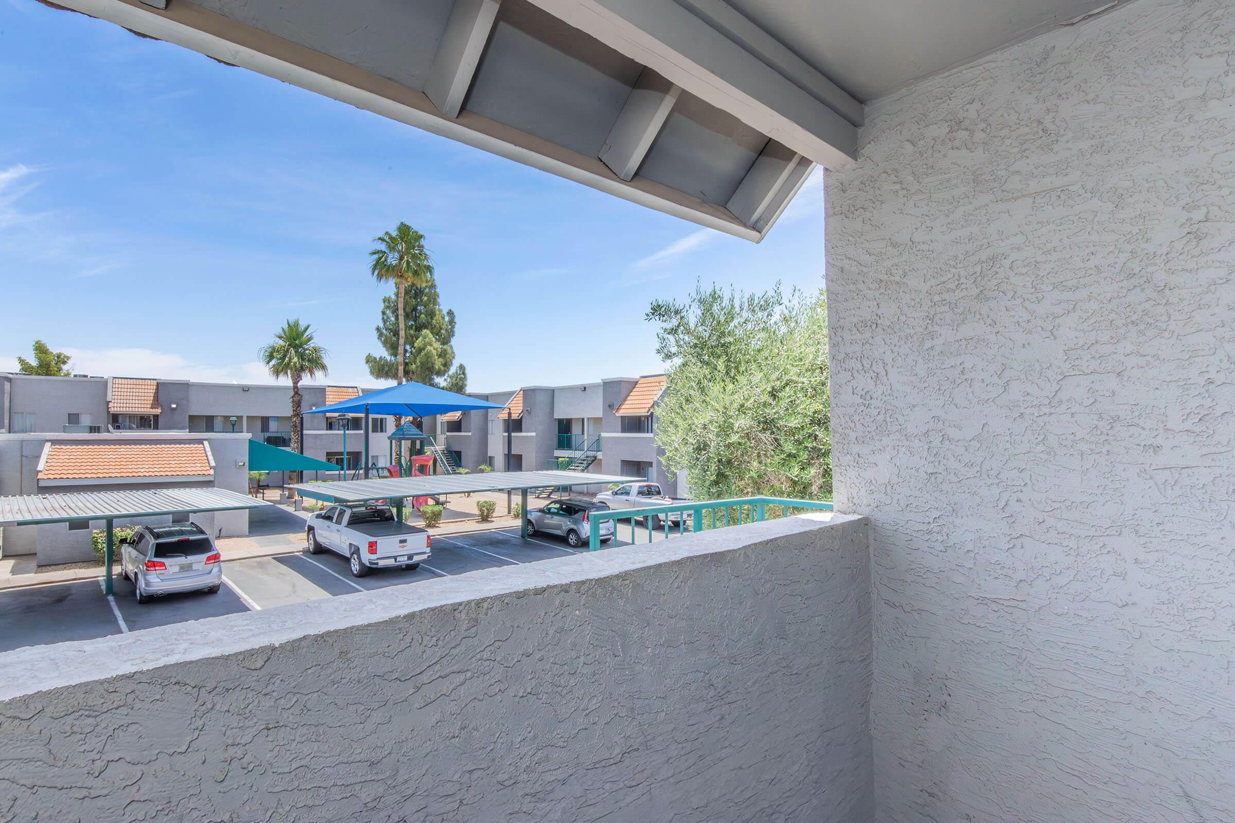 View from a balcony overlooking a residential area. Below, there are parked cars and a shaded area with a blue canopy. Palm trees and other greenery are visible in the background, along with multi-story apartment buildings under a clear blue sky.